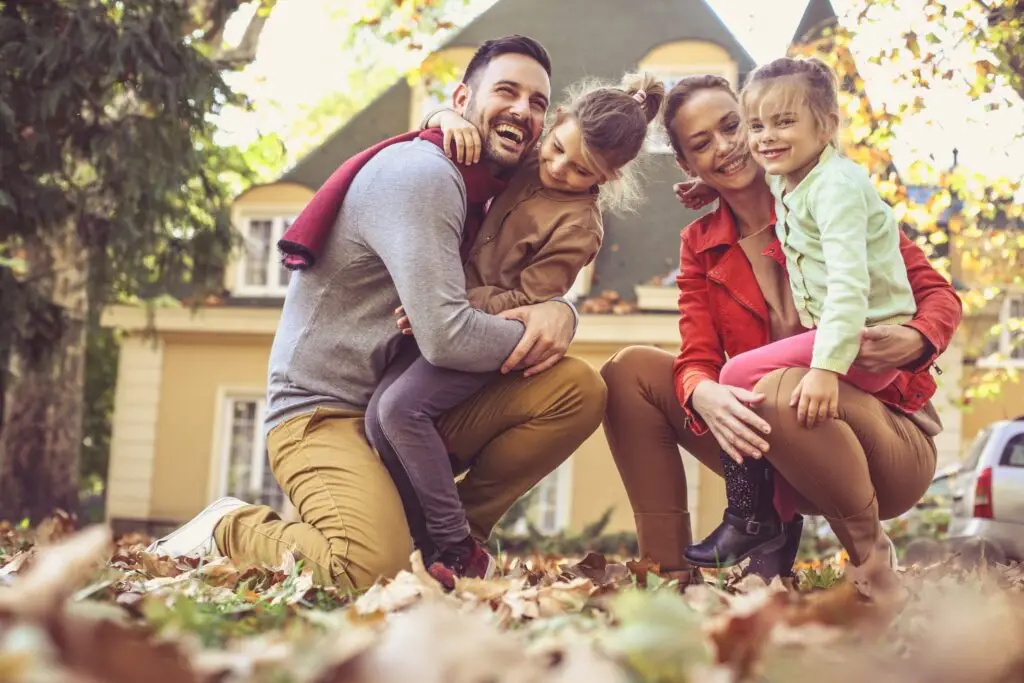 A family of four is joyfully embracing in a leafy yard on an autumn day, as they create great family memories. The parents crouch down to hug their two young children, against a backdrop of a house with a sloping roof and surrounding trees filled with vibrant fall foliage.