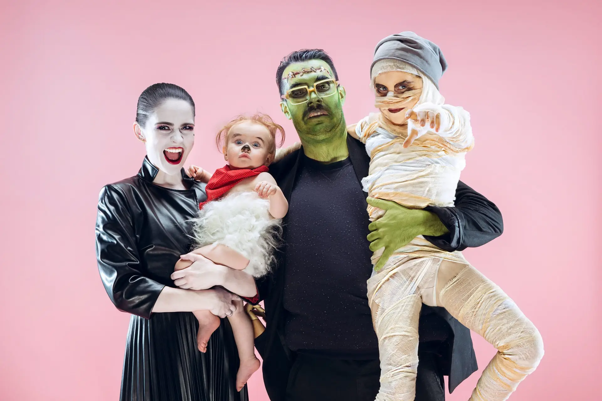 A family dressed in Halloween costumes poses against a pink background, showcasing creative family Halloween costume ideas. The adults are transformed into a vampire and Frankenstein, while the kids charm as a mini sheep and mummy.