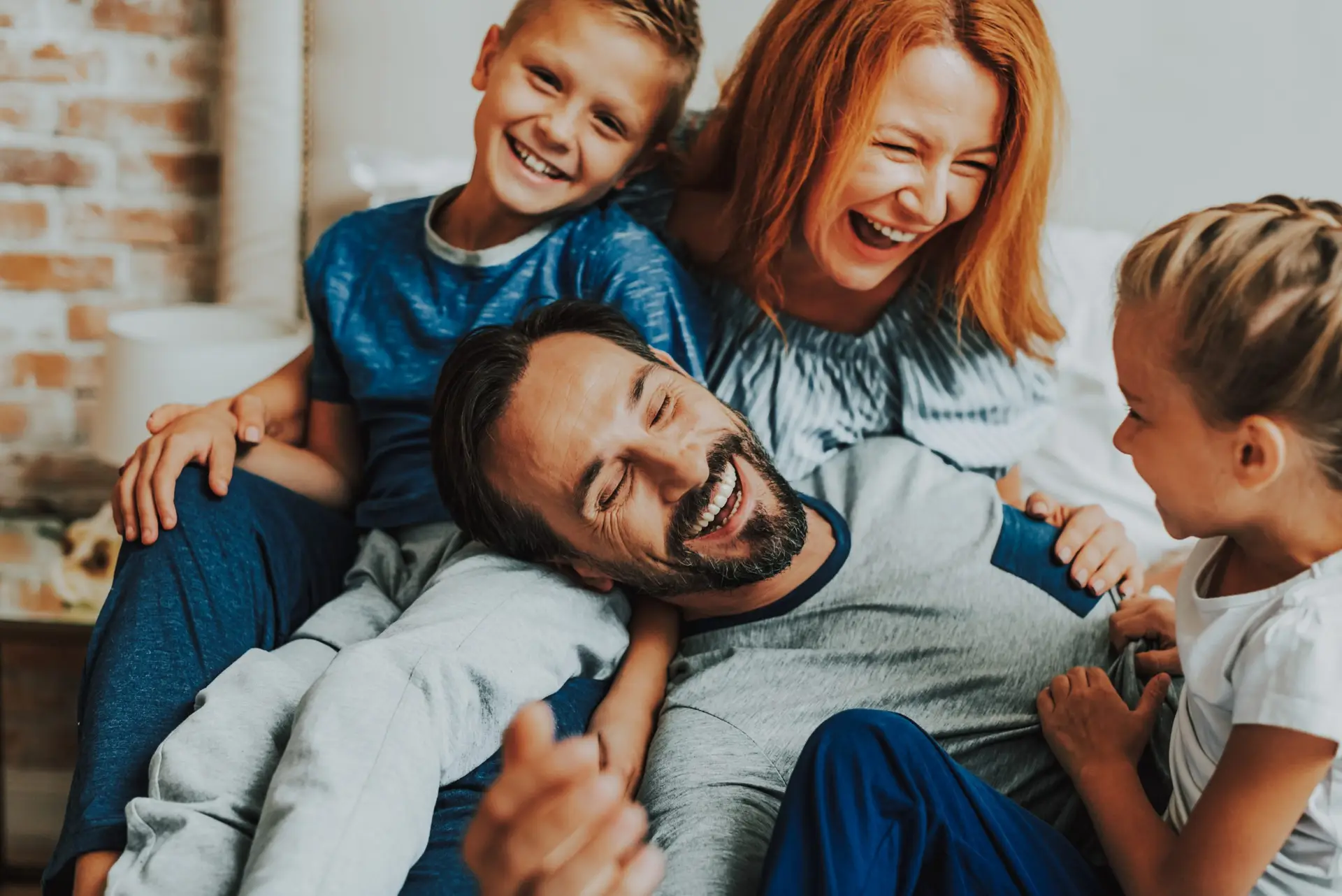 A joyful family of four, including two children, is sitting together on a couch. As they laugh and hug, with the father lying down and being playfully surrounded by his family, everyone appears happy and relaxed—a true example of how to be a role model to your children.