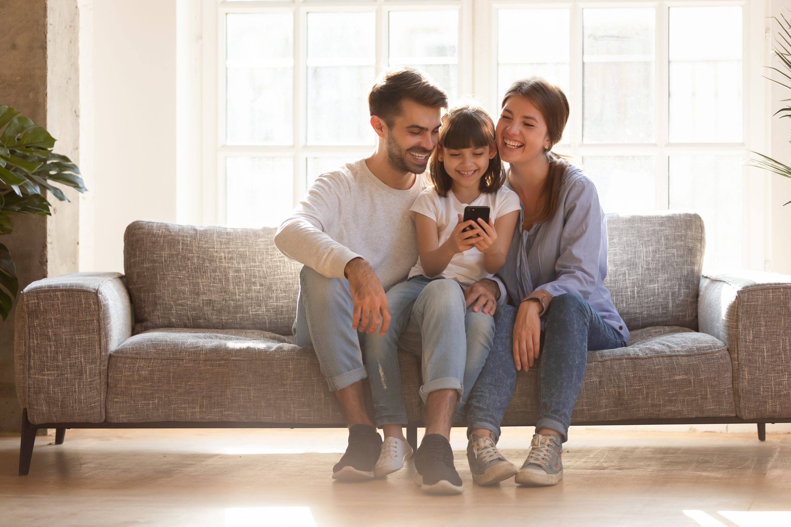 A family of three sits on a couch in a bright living room. The child is holding a smartphone, using the best child control app, smiling as the parents lean in to look at the screen. Their expressions are joyful and relaxed, with sunlight streaming through large windows.