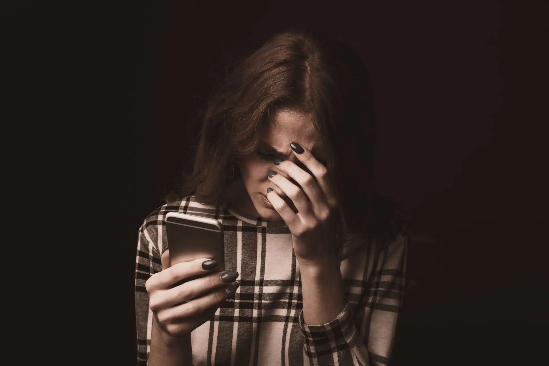 A woman in a plaid shirt looks distressed while holding a smartphone in one hand and covering her face with the other, aware of the dangers of chat rooms. The background is dark, emphasizing her expression and the eerie glow from the device.