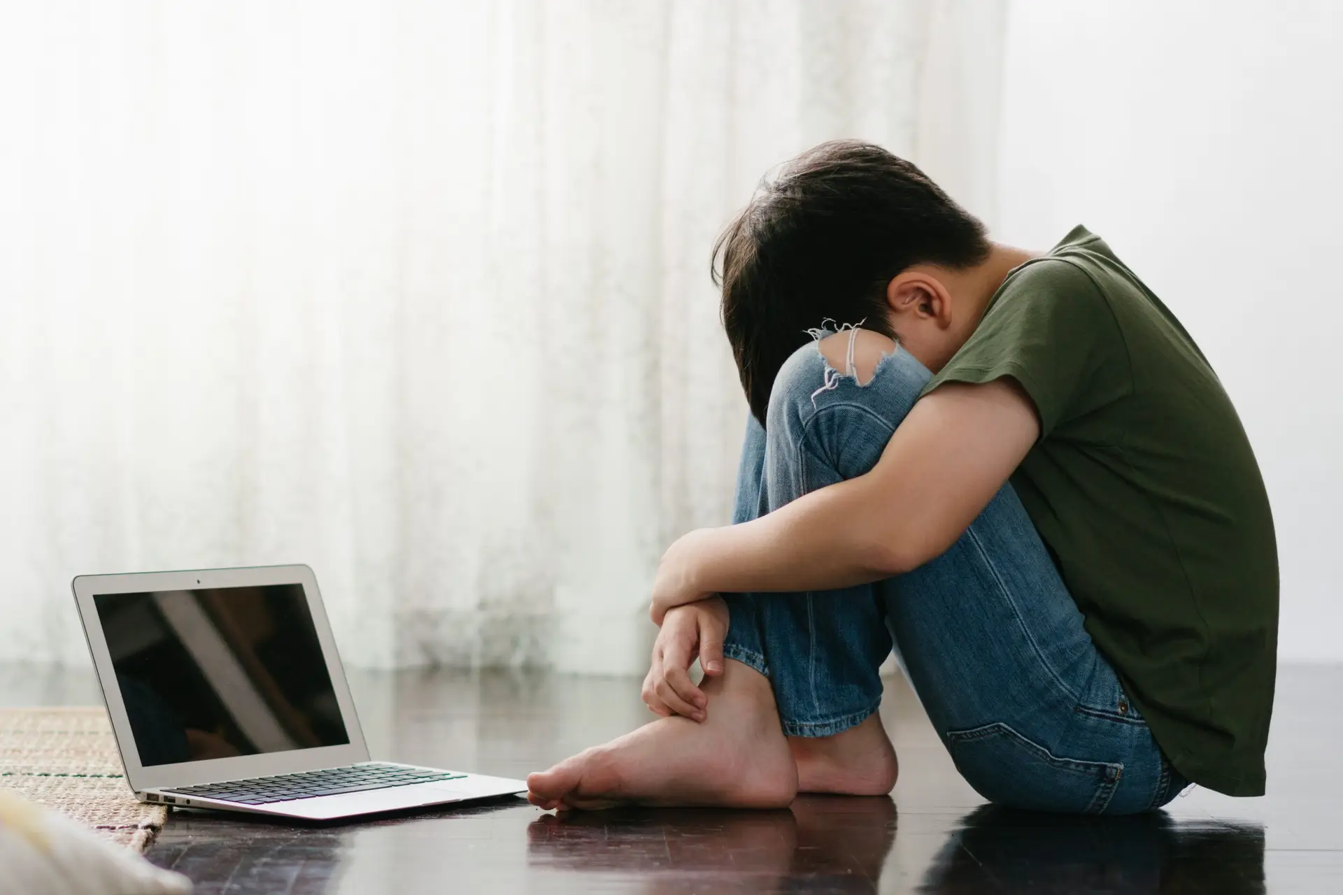 A person sits on the floor with knees pulled to their chest, a hint of fear in their eyes, head resting on their arms next to an open laptop. The room is softly lit with sheer curtains in the background, merging tech with a calm ambiance.