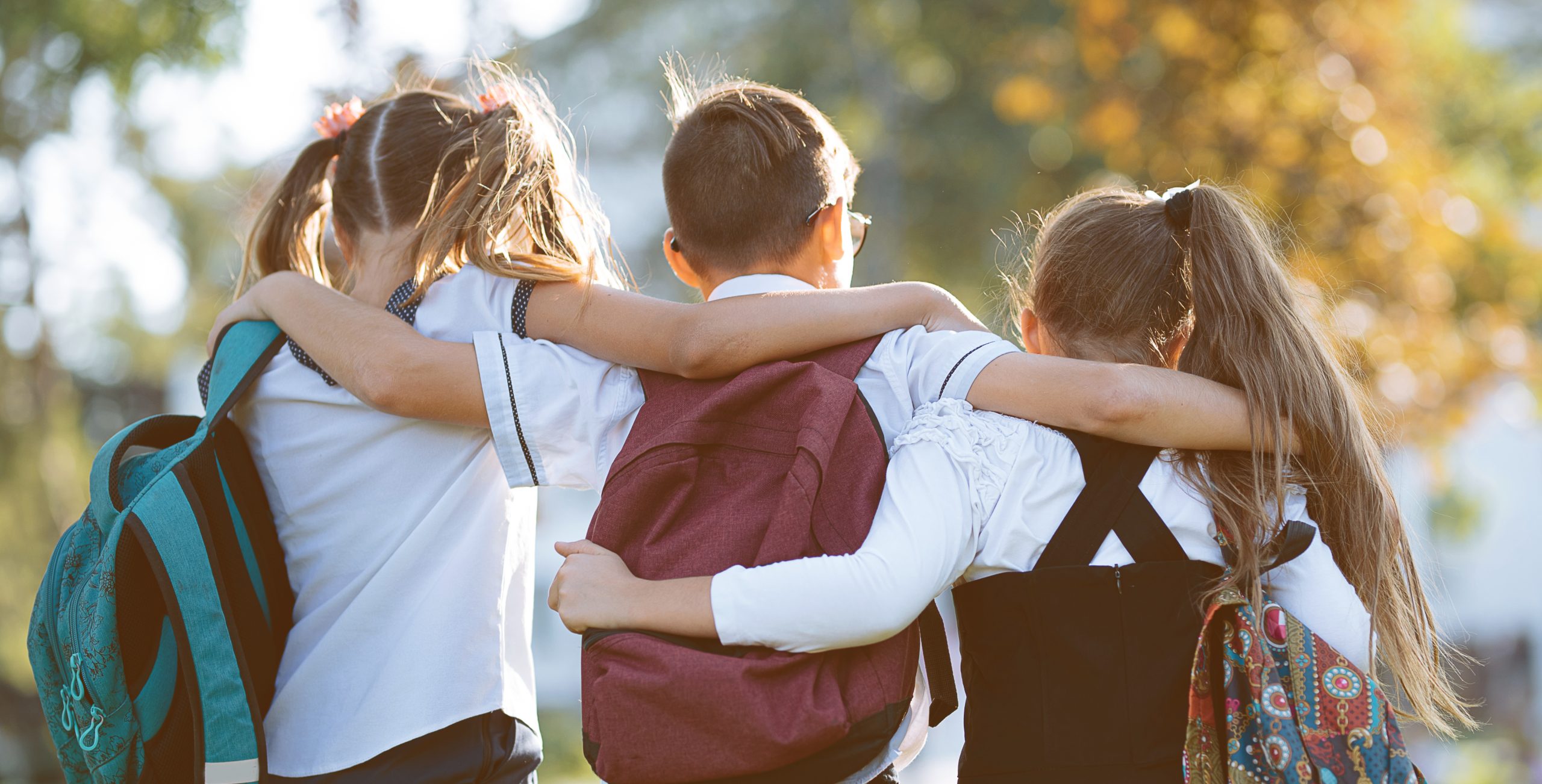 Three children with backpacks walk arm in arm outdoors, embodying friendship and togetherness. As the sun shines through the trees, creating a warm atmosphere, they are not only enjoying nature but also learning about teaching boundaries to youth in an organic way.