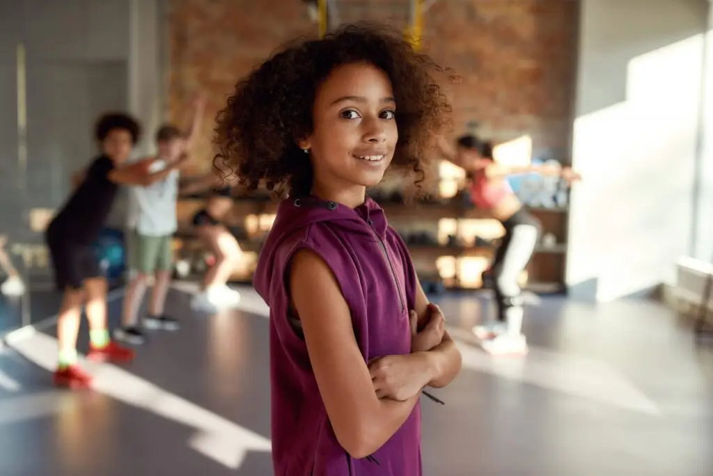 A young girl with curly hair in a purple sleeveless top stands confidently with arms crossed in a gym, where a group of kids exercises in the background. Sunlight streams through large windows, reminding us why sleep and nutrition are so important for teens to fuel their active lives.