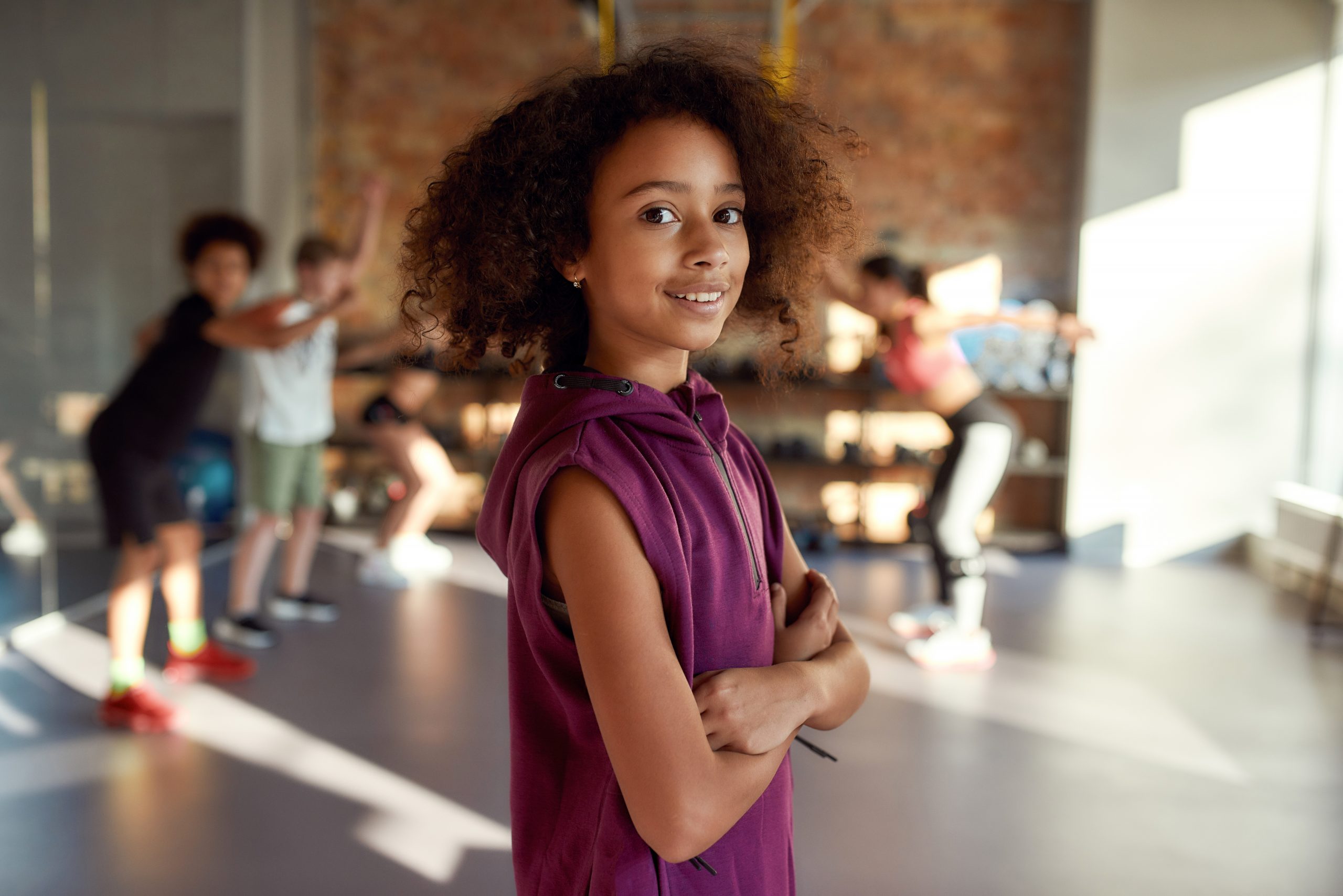 A young girl with curly hair in a purple sleeveless top stands confidently with arms crossed in a gym, where a group of kids exercises in the background. Sunlight streams through large windows, reminding us why sleep and nutrition are so important for teens to fuel their active lives.