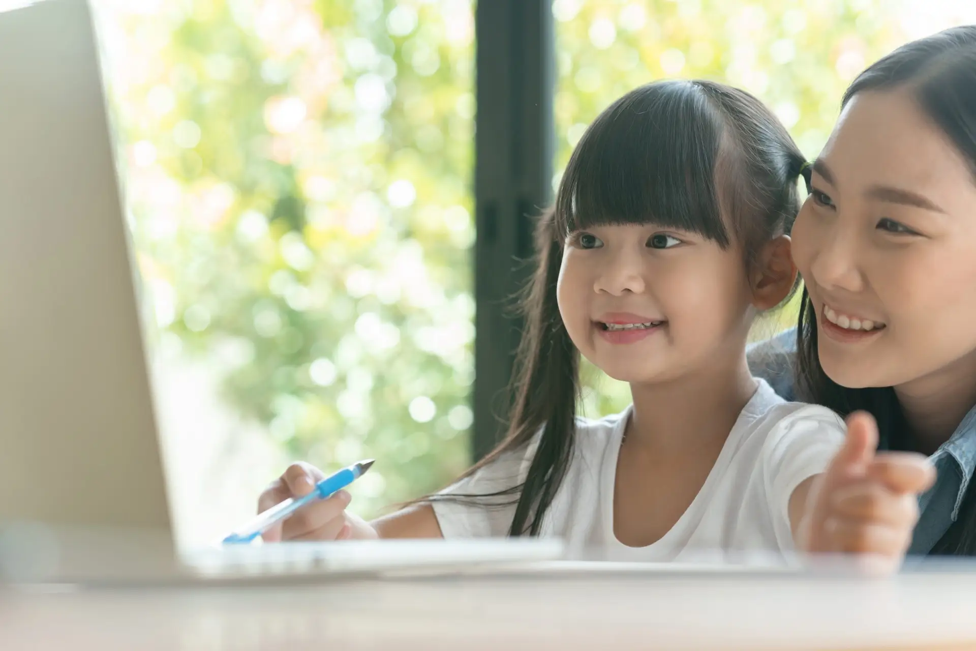 A mother and daughter are smiling while looking at a laptop screen, exploring how to be kind online. The young girl holds a pen, suggesting they are working or learning together. Sunlight filters through the window, creating a warm, cheerful atmosphere.