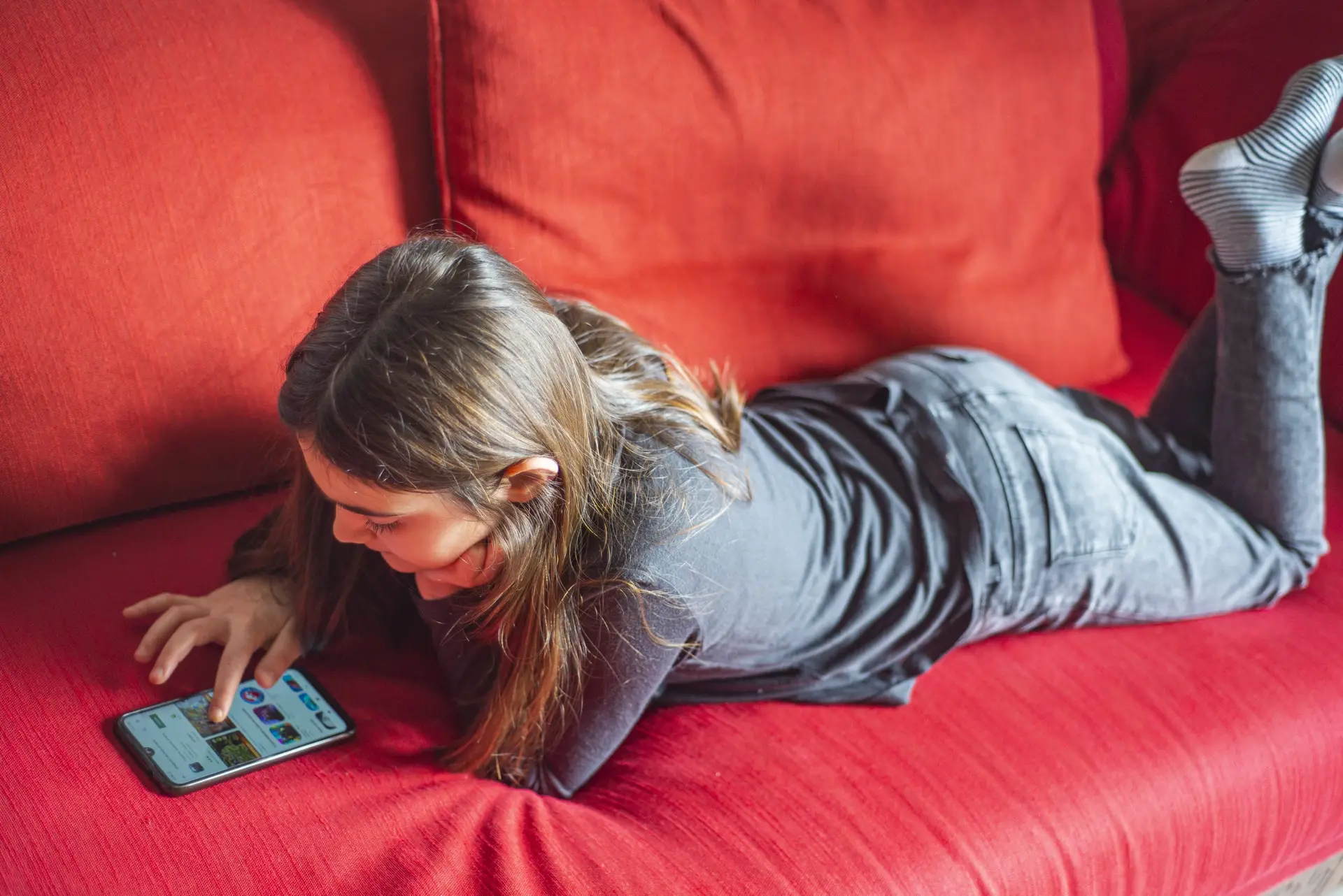 A young girl with long hair lies on a red couch, engrossed in her smartphone. Dressed in a dark outfit, she’s focused on the screen—perhaps exploring a complete guide on dangerous apps for kids—as her feet rest comfortably on the couch's cushions.