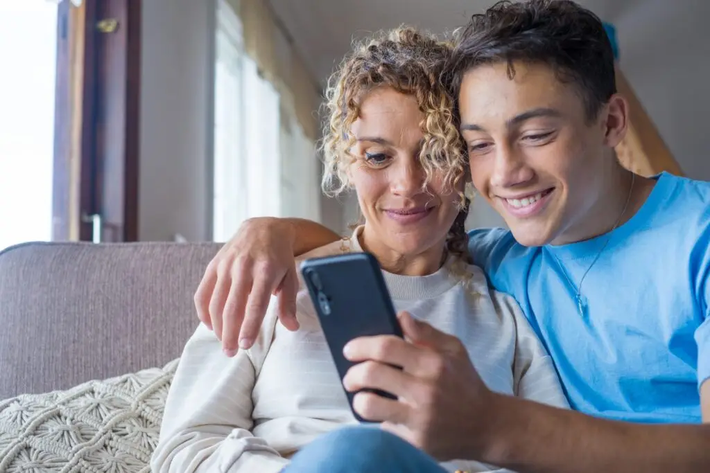 A curly-haired woman and a young man are sitting on a couch, smiling as they look at a smartphone together, enjoying the freedom their flexible schedules allow. The room is softly lit with natural light filtering through a window in the background.