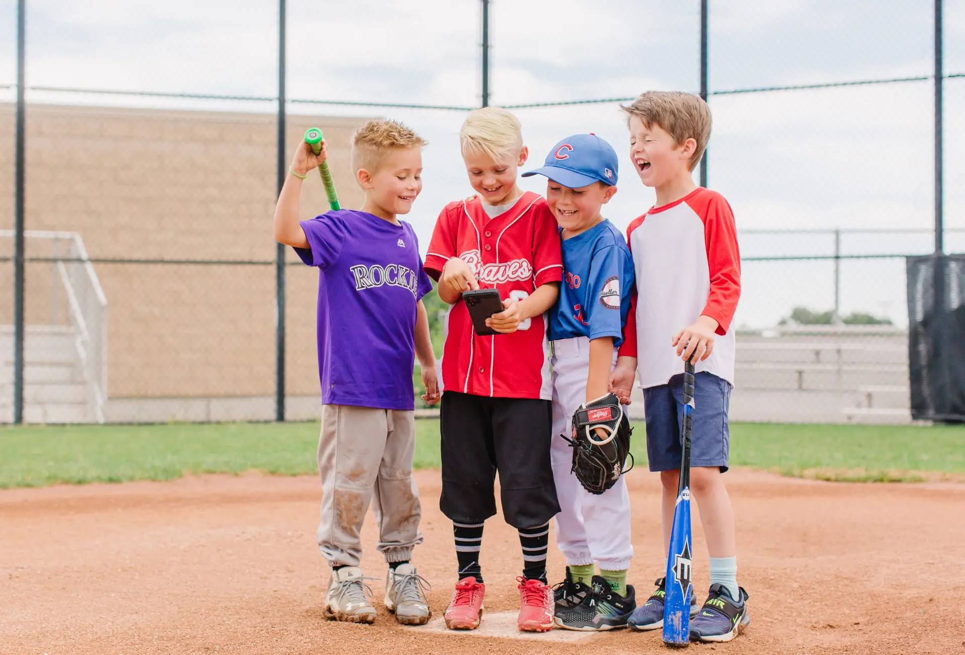 Four young boys in baseball attire stand together on a field, smiling at a smartphone held by one of them. Nearby, a blue baseball bat and glove rest on the ground, surrounded by a chain-link fence and grassy area. They embody sport safety for kids, reminding parents what to prioritize on the field.