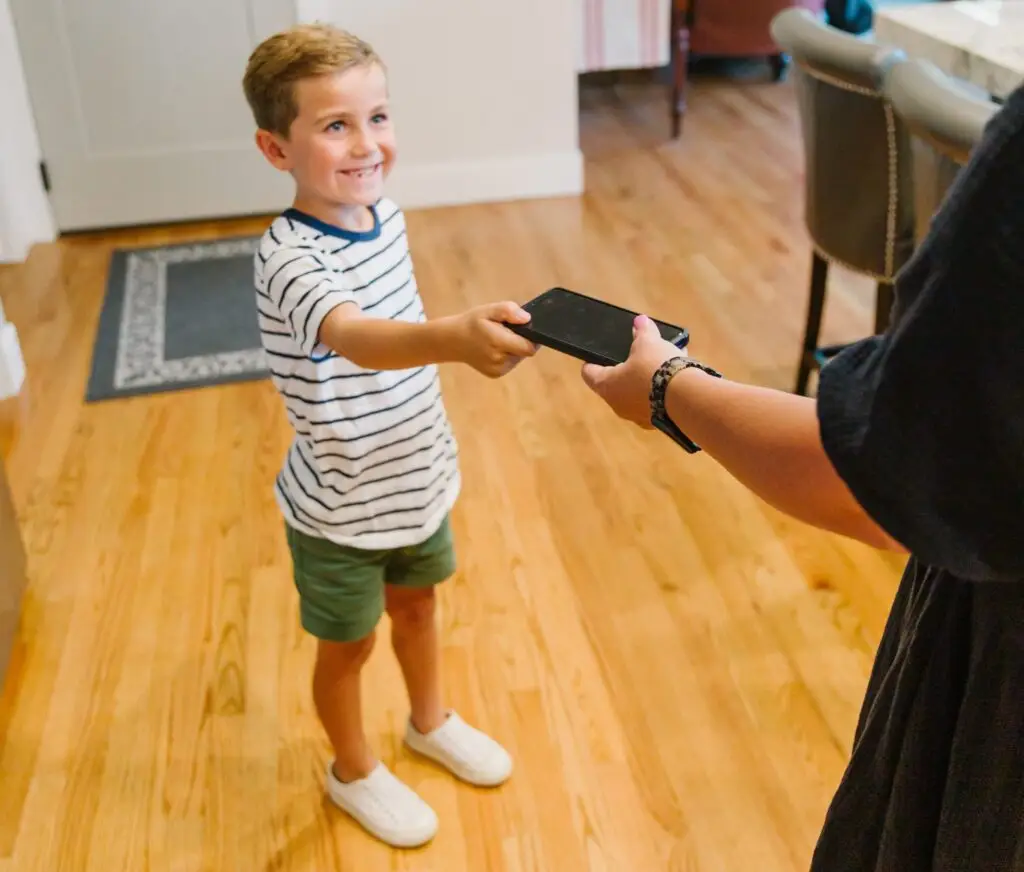 A young boy wearing a striped shirt and green shorts smiles as he confidently hands a phone to an adult in a black outfit. They stand in a room with wooden floors and a carpet near a white door, suggesting there's no need to be afraid of giving your child a phone.