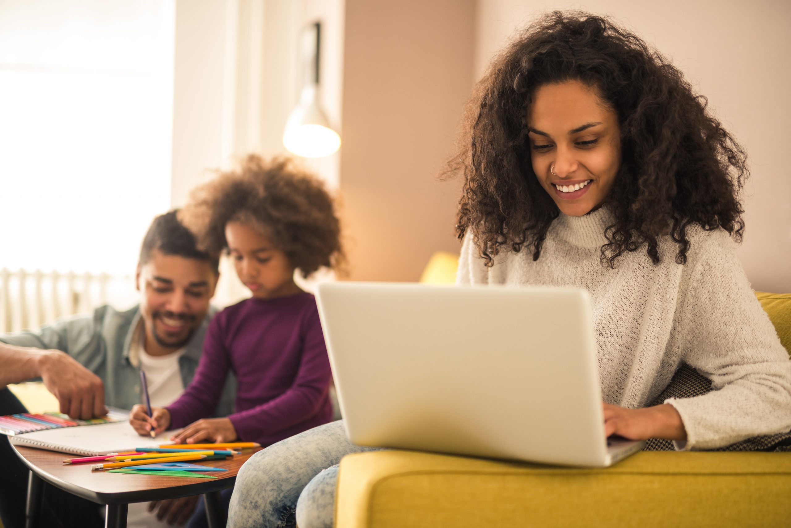 A woman sits on a yellow couch working on a laptop, smiling as she scrolls through "Your Guide to Managing Cookies." Nearby, a man and young girl color with crayons at the table, creating a cozy family scene in the living room.