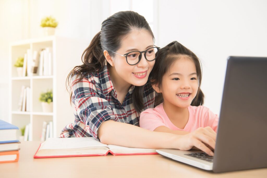 A woman and a young girl smile while looking at a laptop screen, perhaps discovering tips on how to learn to type faster. The woman sports glasses and a plaid shirt; the girl dons pink. An open notebook lies beside them in the bright room, capturing their shared curiosity.