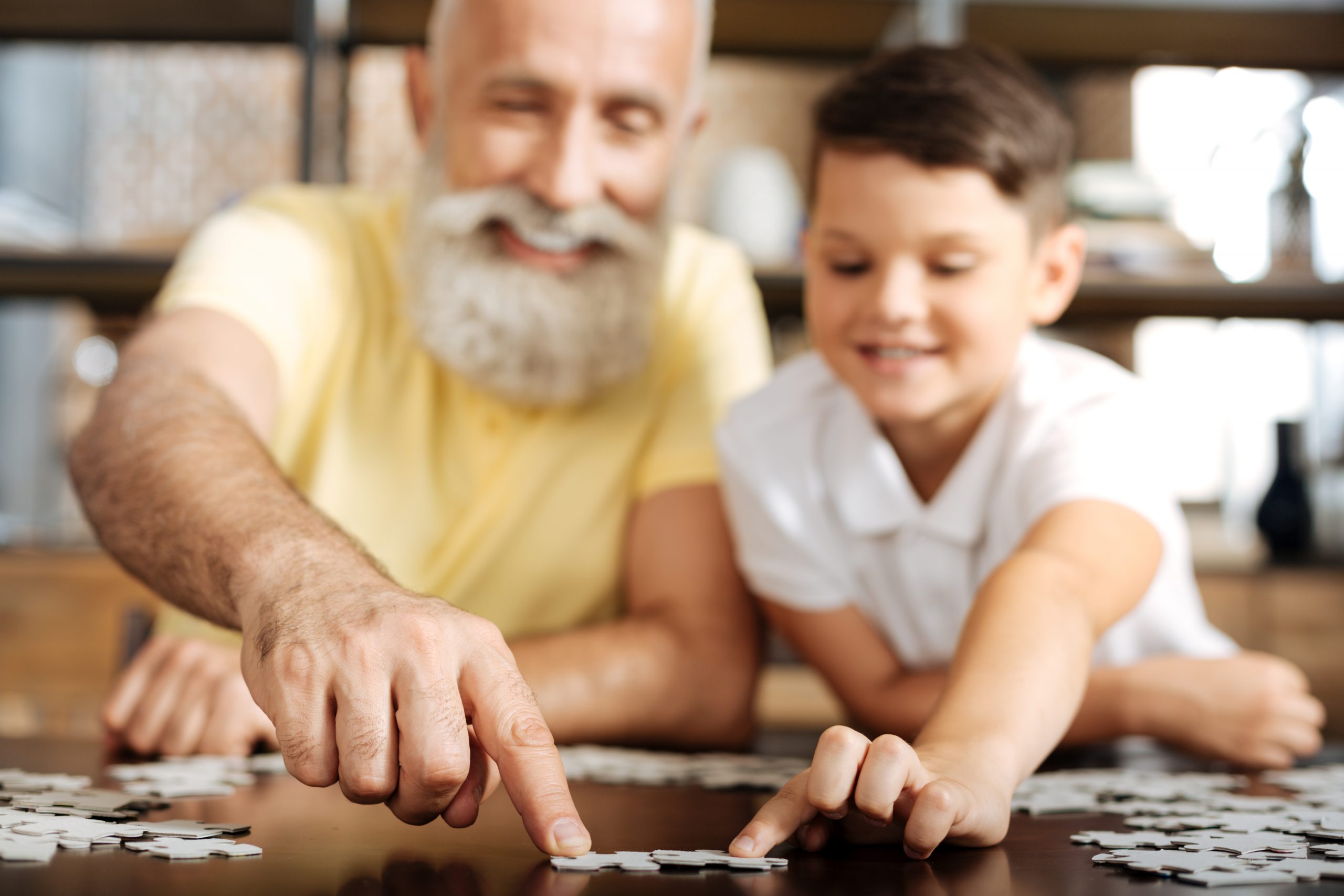 An elderly man with a beard and a young boy are smiling as they experience the joy of assembling a jigsaw puzzle together on a table. The softly blurred background highlights their focus, showcasing one of the ten benefits of puzzles at every age: bonding across generations.