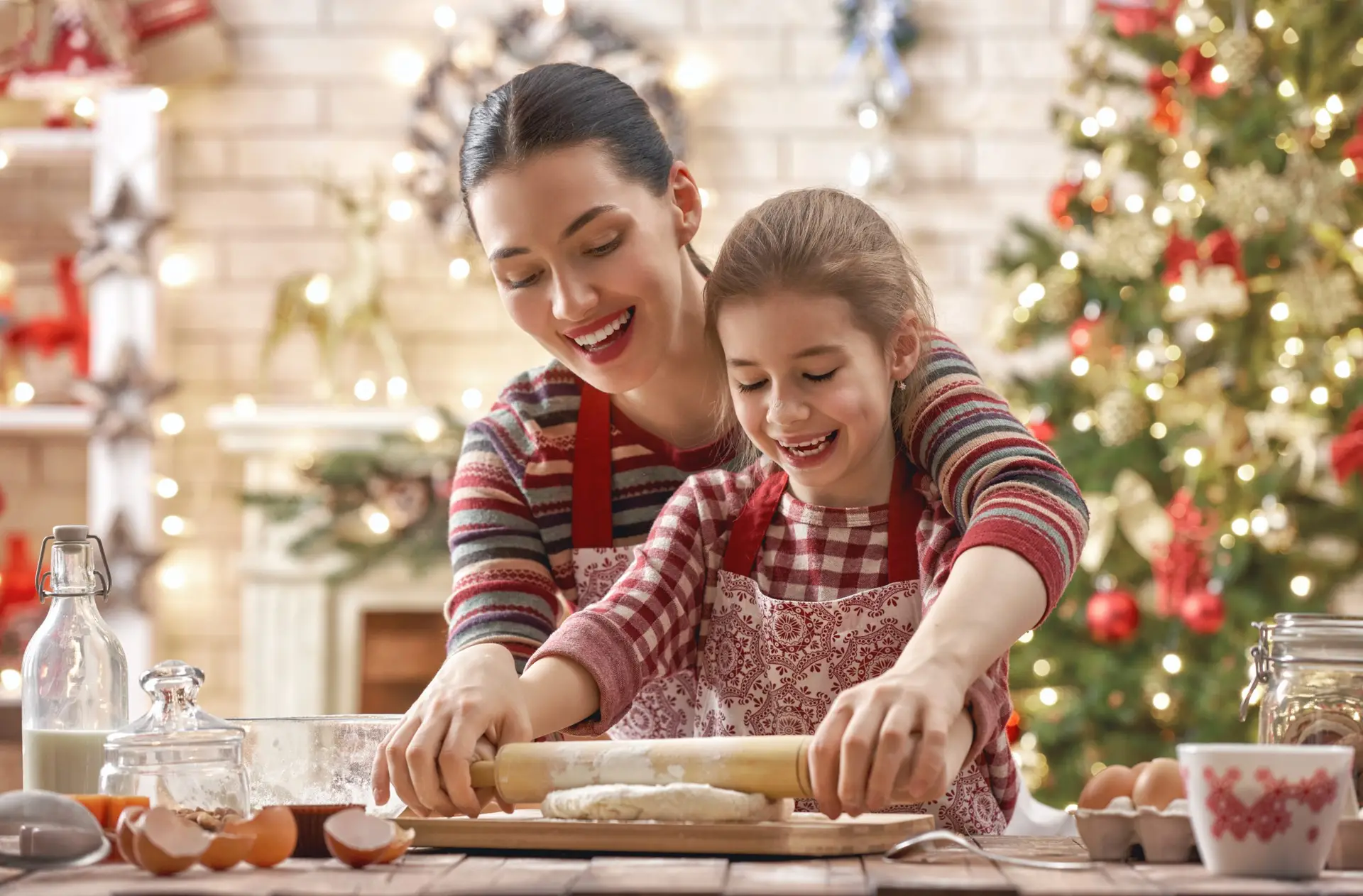 A woman and a young girl, both in striped shirts and aprons, smile while rolling out dough together. In their cozy, festive kitchen adorned with holiday lights and a Christmas tree, they embrace the joy of A Christmas without Screens. Every parent should try this magical experience!