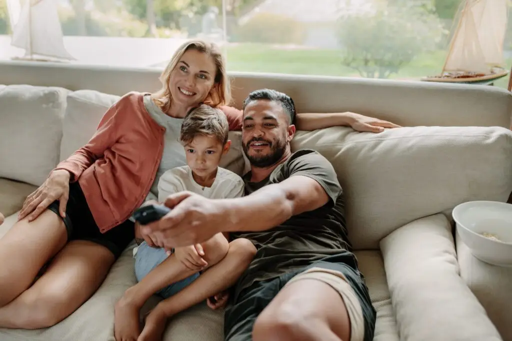 In a cozy living room, a family of three relaxes on the sofa, enjoying time together. Soft furnishings and greenery through the large window add to their serene moment, as if they’ve stepped out of a scene where reality TV shows might ponder if they do background checks on such warmth.