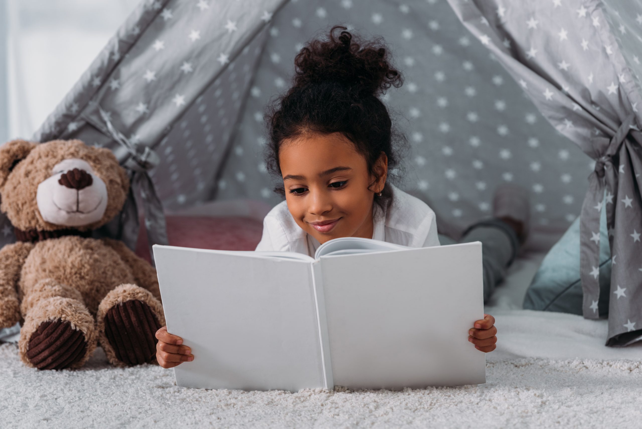 A child lies on their stomach, engrossed in a book under a gray star-patterned tent, one of Five Ways to Wind Down Without Your Phone. A plush teddy bear keeps them company on the soft, carpeted floor.