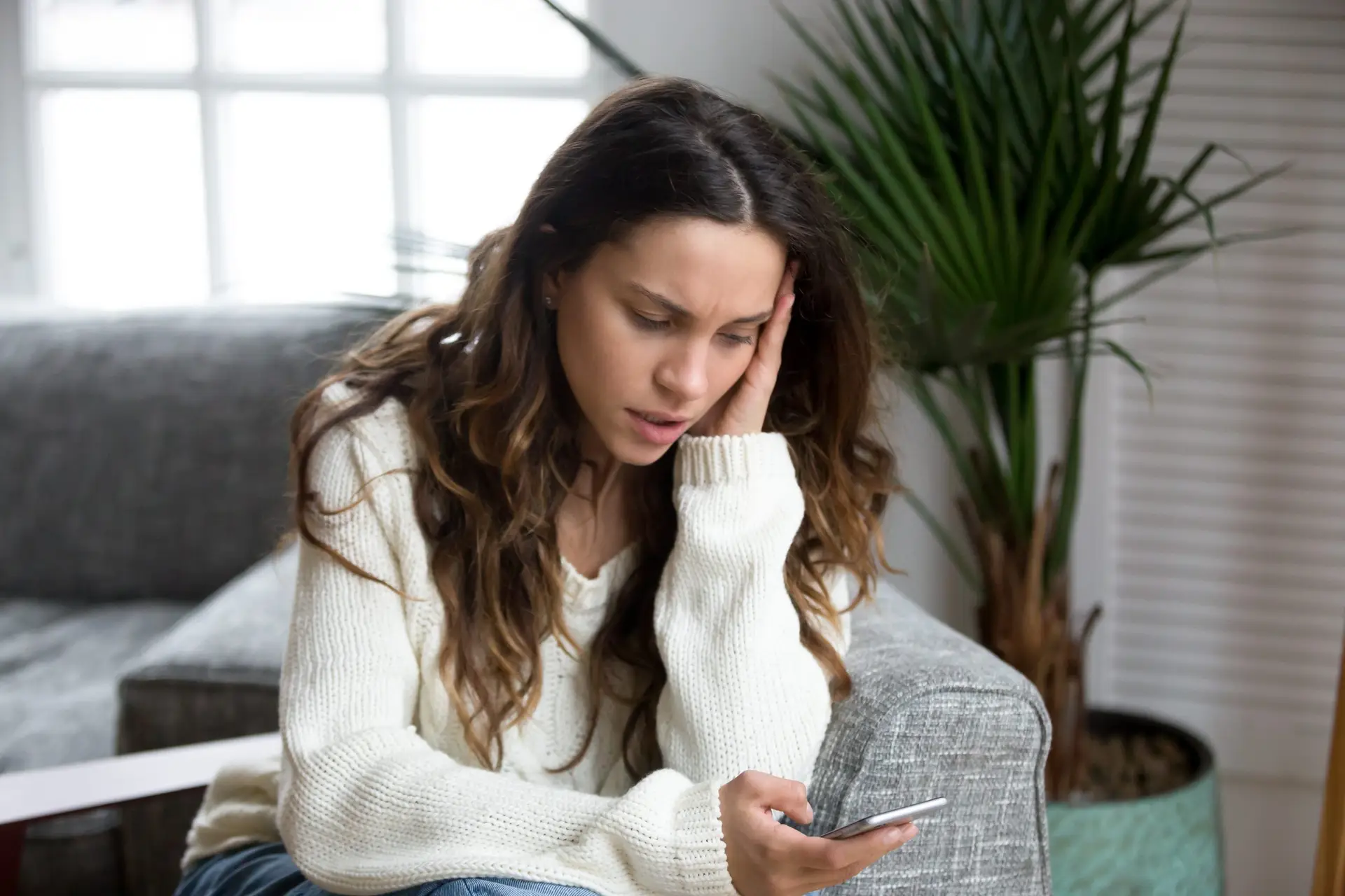 A woman with long hair, wearing a white sweater, sits on a couch with a concerned look, holding her smartphone. She's the mother of three who bravely poses as a teen girl online to expose sexual predators. A potted plant is in the background near a window.