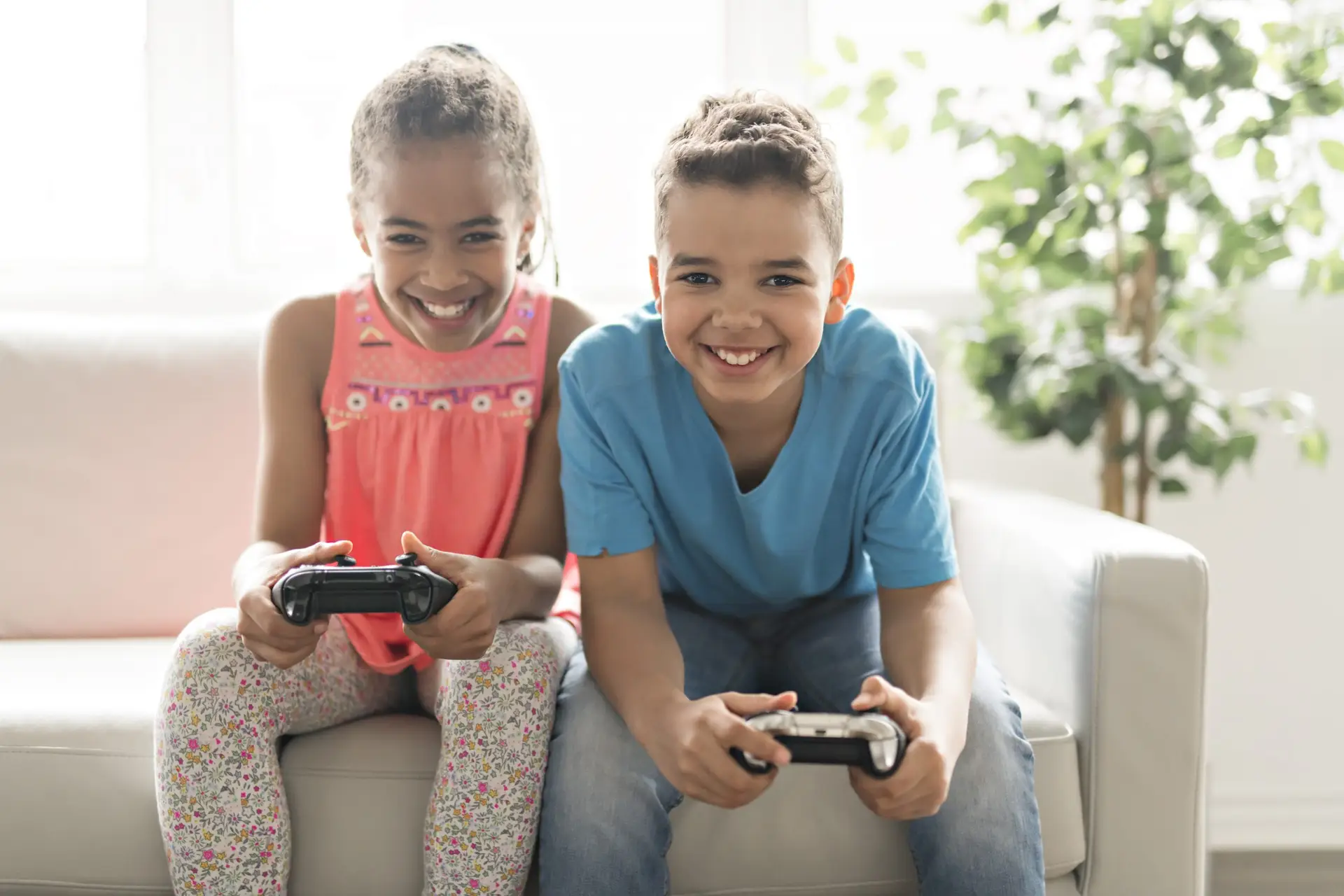 Two children sit on a couch, smiling and immersed in video games. The girl on the left wears a pink top with patterned leggings, while the boy on the right sports a blue shirt with jeans. A houseplant adds charm to their cozy setup, where they could likely explore 7 tips for good gaming etiquette with friends.