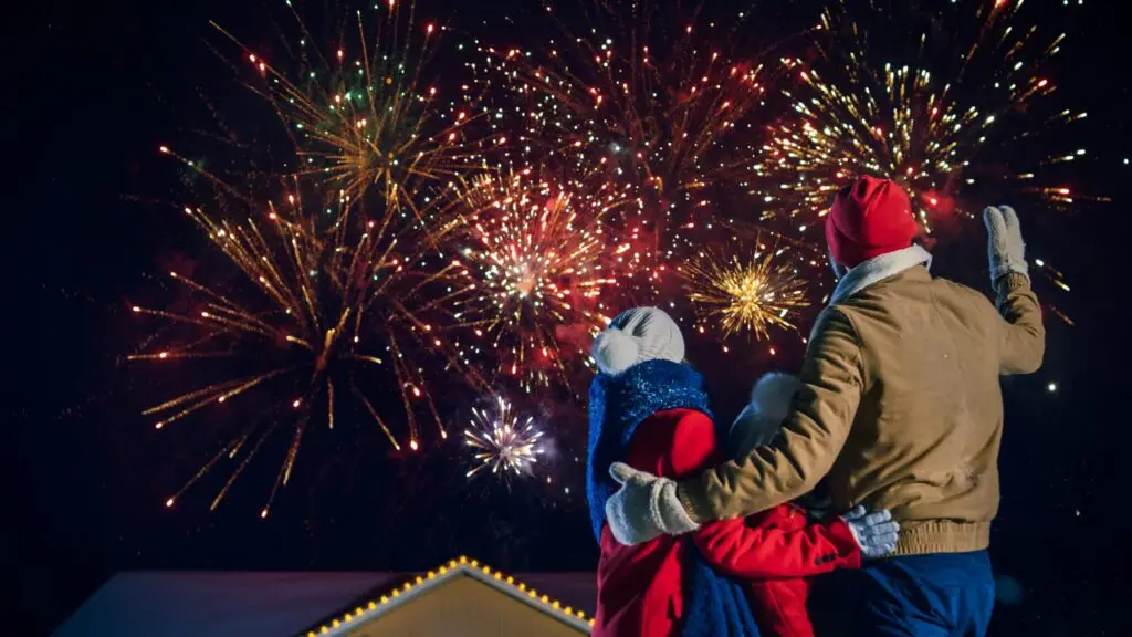 A family of three bundled in winter clothing stands outside at night, watching vibrant fireworks explode safely in the sky. The scene is festive, with colorful bursts of light illuminating the dark background above a house with glowing roof lights. Parents keep firework safety tips in mind for a joyful celebration.