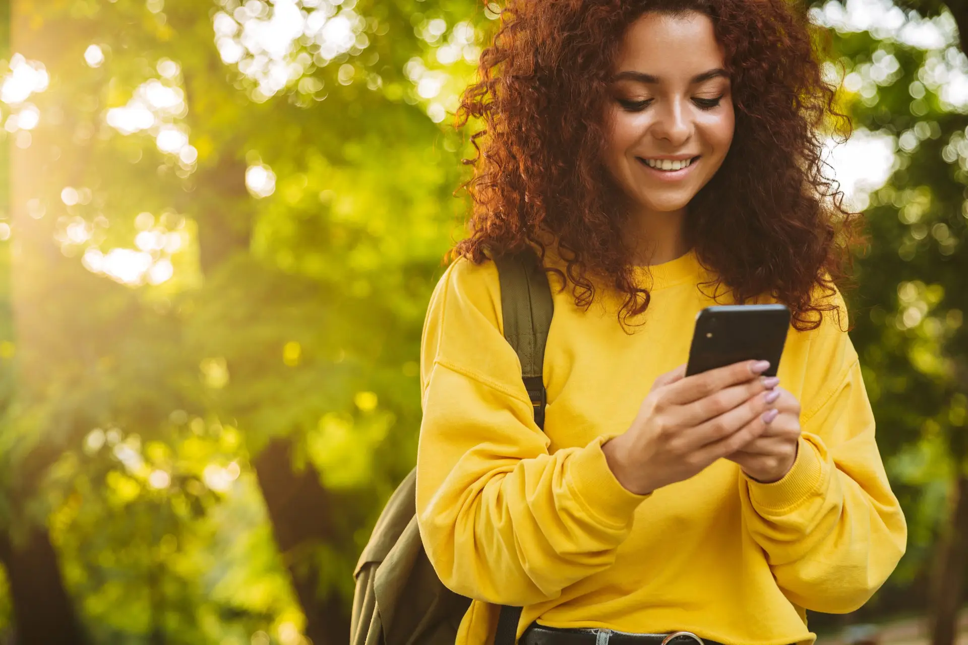 A young woman with curly hair, wearing a yellow sweater and backpack, smiles while looking at her phone. She stands outdoors, surrounded by lush green trees and sunlight filtering through the leaves, perhaps pondering phone and texting etiquette for better communication.