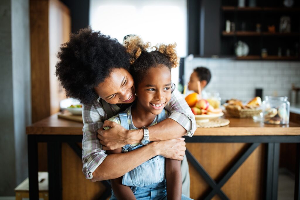 A parent hugs their smiling child from behind in a warmly lit kitchen, imparting values and morals in a digital world. A table with fruit and baked goods is behind them, while another child is visible in the background, learning through love and guidance.