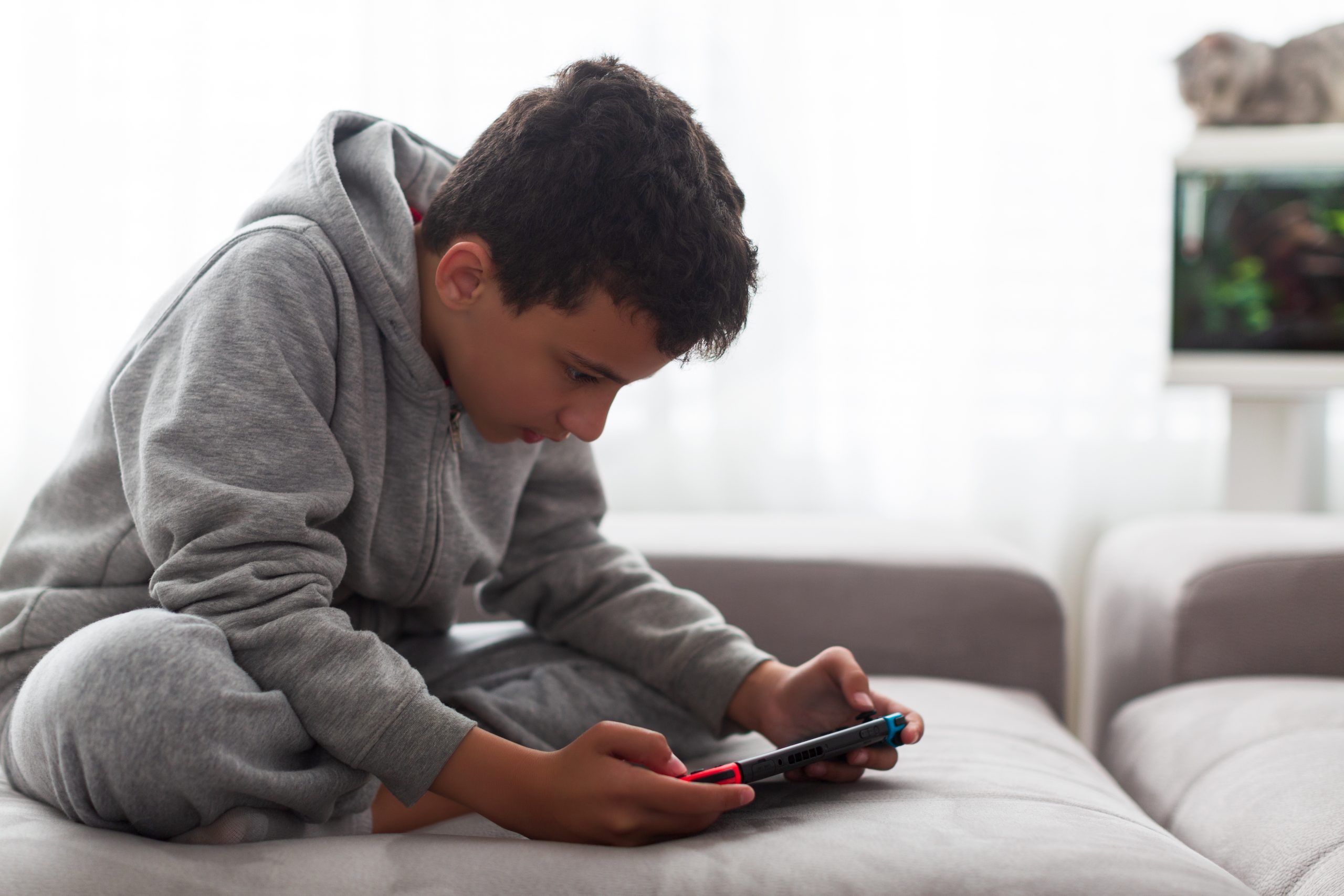 A young boy in a gray hoodie sits cross-legged on a light-colored couch, intently engaged with a handheld gaming console. The room seems bright with natural light streaming through sheer curtains, prompting thoughts like "How can I protect my child from devices?