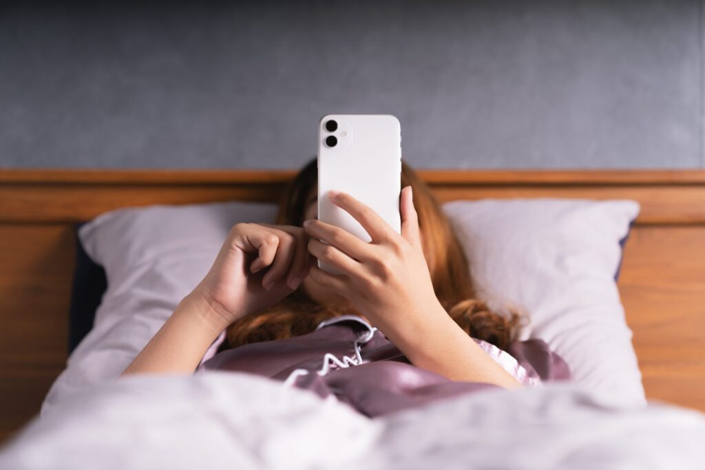 A person lying in bed is holding a smartphone above their face, unaware of the advice "don't sleep with your phone." They are under a blanket, with a wood-paneled headboard and gray pillows in the background.