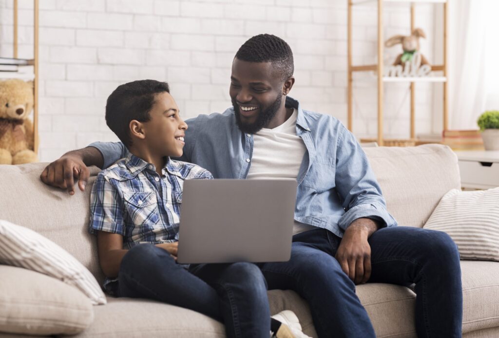 A man and a boy sit on a couch with a laptop, smiling at each other as they explore tech together without being controlling. The man has his arm around the boy in their cozy room decorated with a teddy bear and plant-filled shelves, fostering trust and understanding.