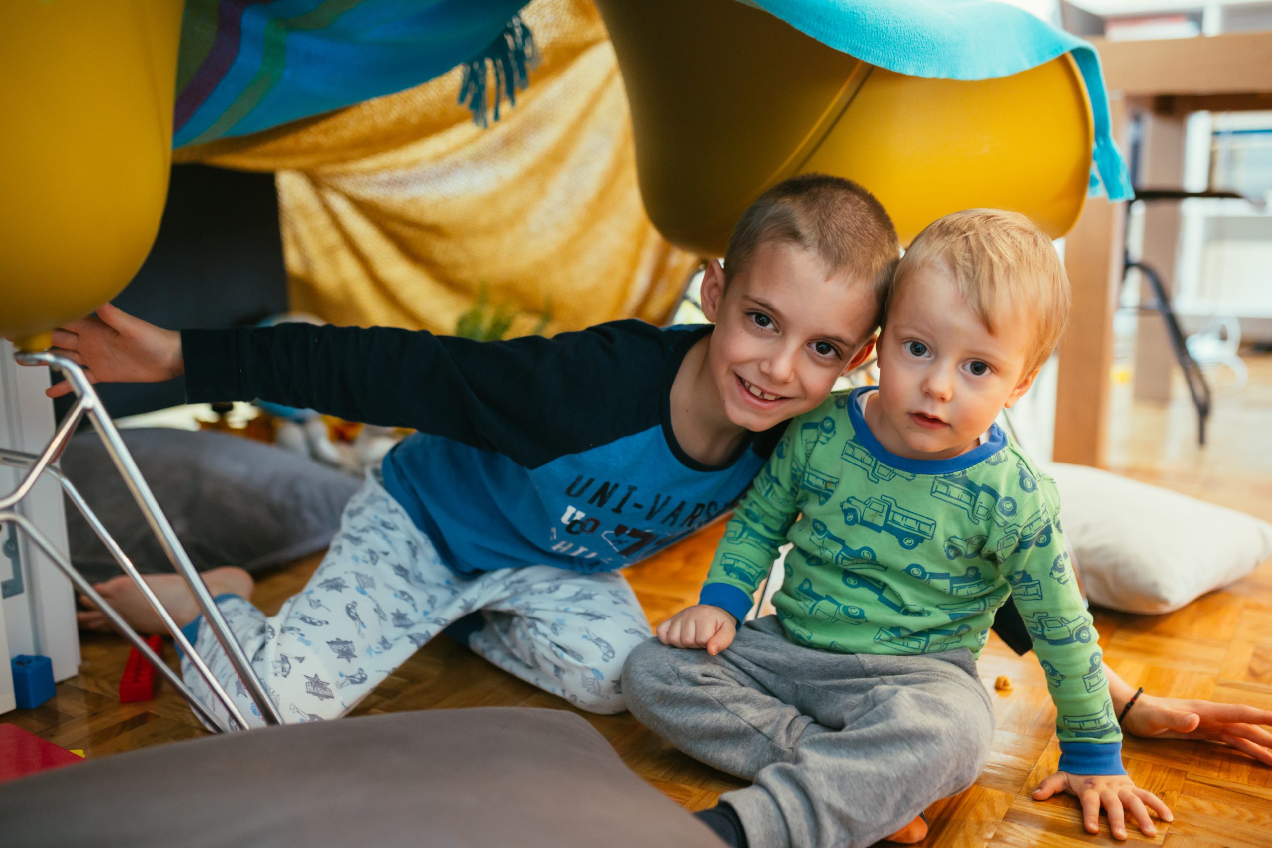 Two young children sit together under a blanket fort made from cushions and a chair. The older child, wearing a blue shirt, smiles while hugging the younger child, who is in green pajamas. As they enjoy their cozy indoor setting, they discuss the ten benefits of puzzles at every age.