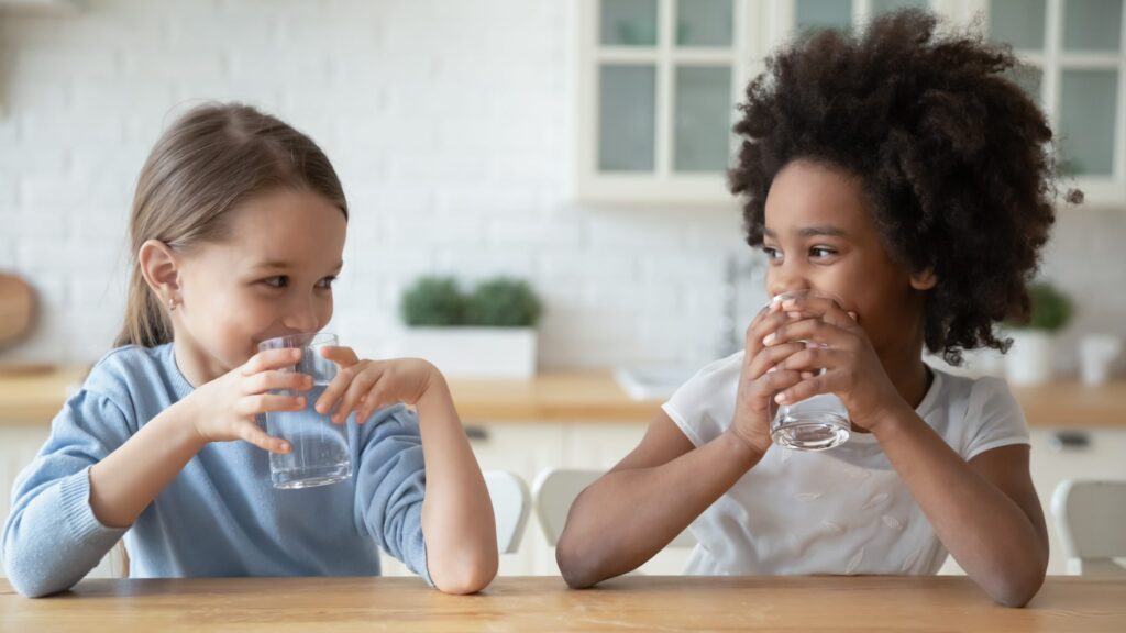 Two young girls sitting at a table, smiling at each other while holding glasses of water in a bright kitchen setting. One has straight hair, and the other has curly hair. The background shows blurred kitchen cabinets and plants.