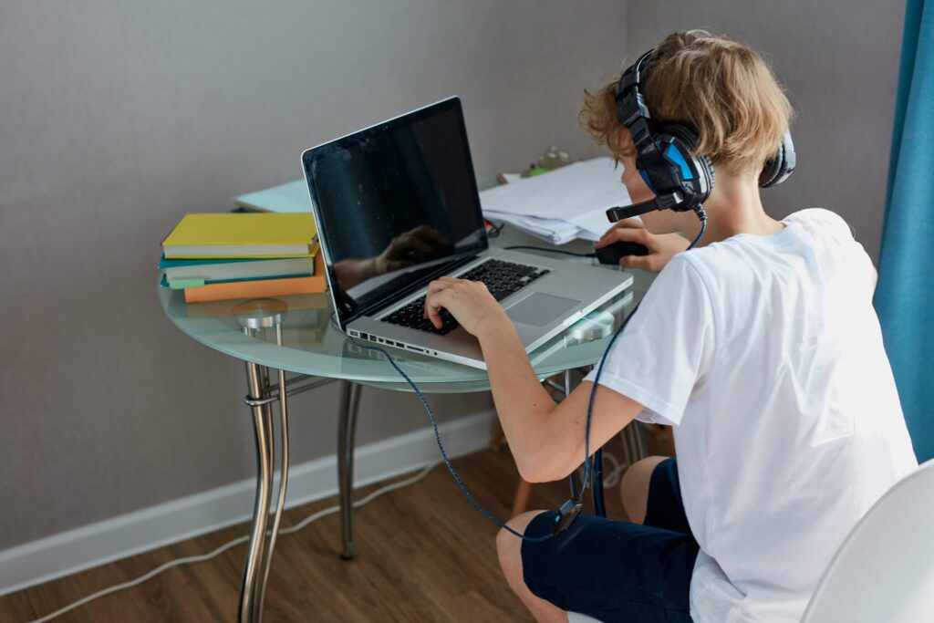 A person wearing headphones uses a laptop on a glass table, exemplifying how to set up an effective learning space. Several books and papers are neatly arranged, fostering focus in the light-colored room with wooden flooring, perfect for kids' home study sessions.