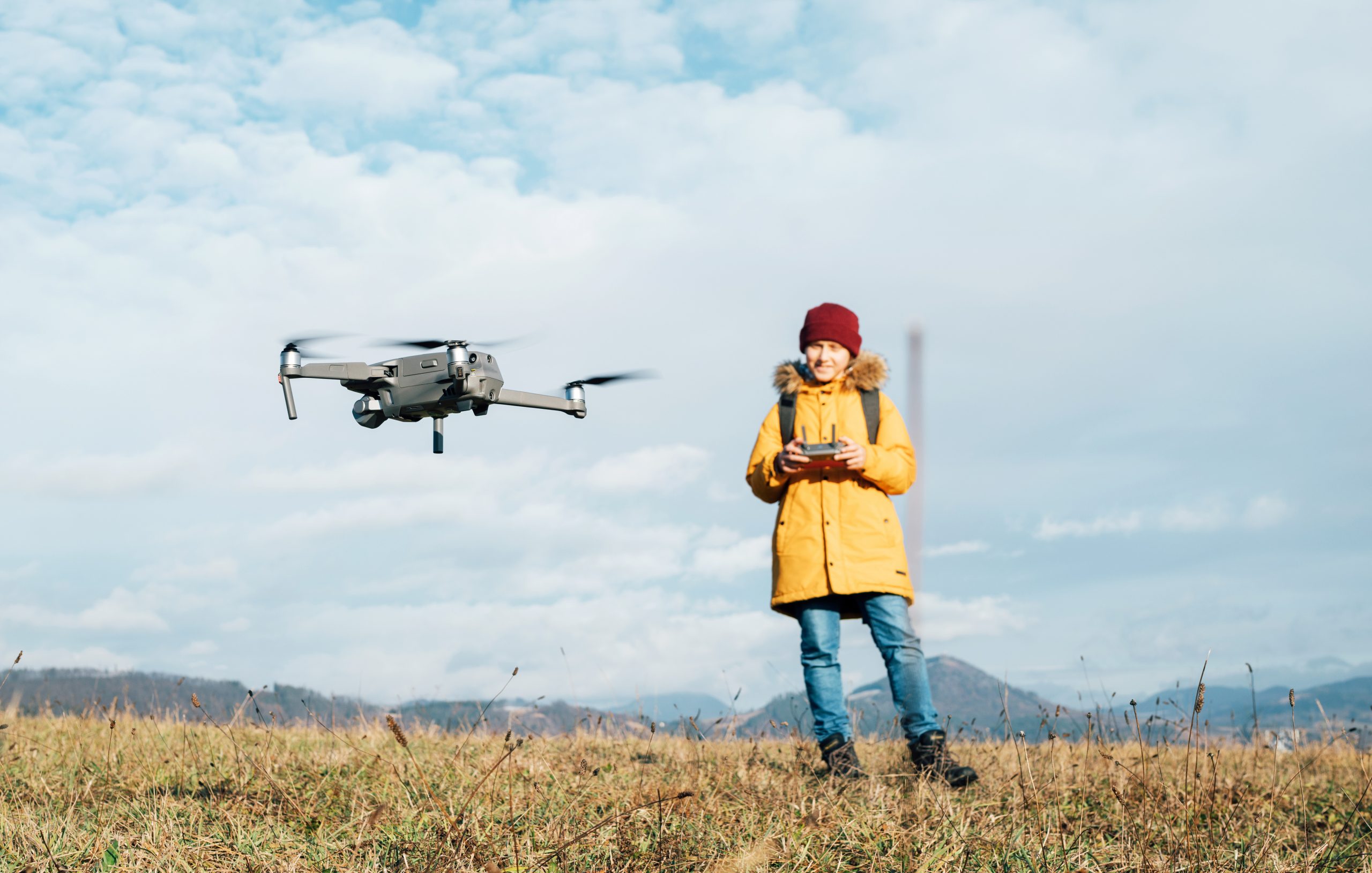 A person in a yellow coat and red beanie operates a drone using a remote control in an open field, embodying the spirit of recreational drone flying. The sky is partly cloudy, and mountains are visible in the distance, combining technology with nature for this beginning pilot's guide.