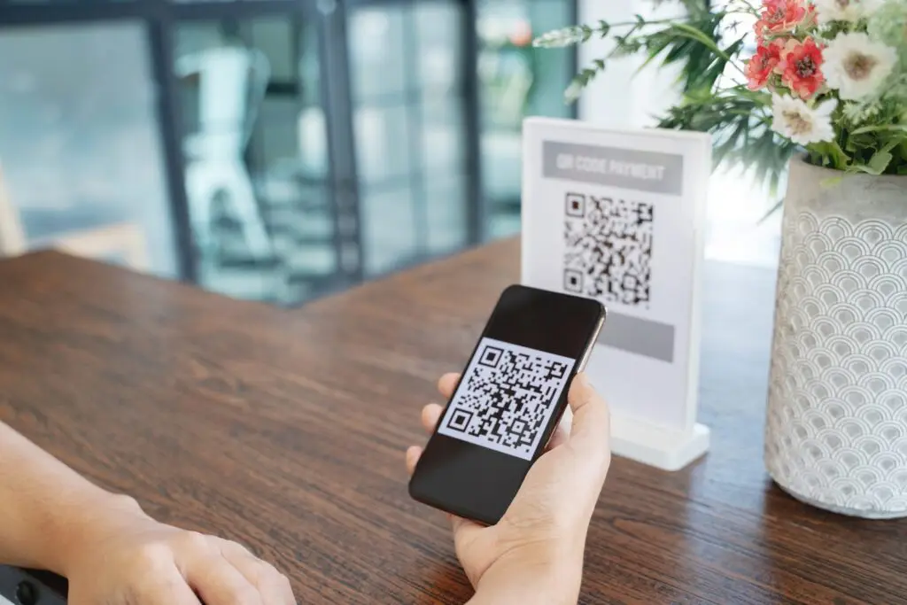 A person holds a smartphone over a table, seamlessly scanning a QR code for payment displayed on a sign. In this modern establishment, with its decorative vase of flowers in the background, this process perfectly exemplifies how intuitive and convenient QR codes have become in our daily transactions.