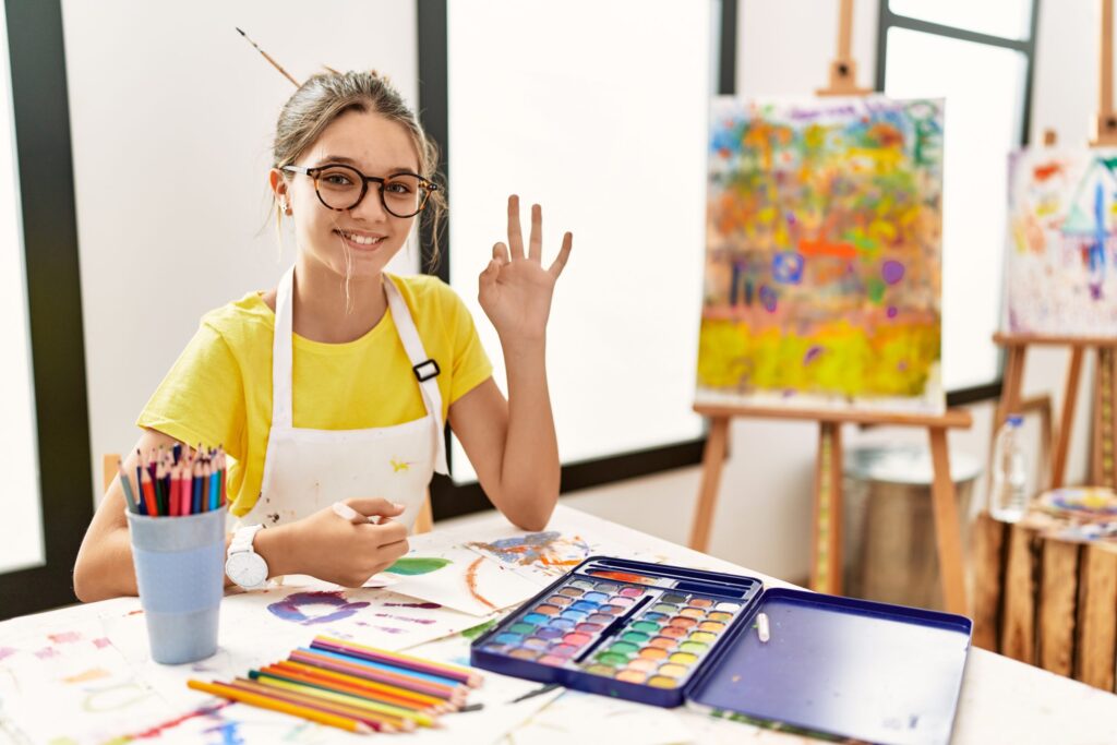 A young girl wearing glasses and an apron sits at a table covered in art supplies, including colored pencils and a watercolor palette. Smiling, she makes an OK hand gesture. Behind her, a colorful painted canvas rests on an easel—a budding artist dreaming of her own Etsy shop.