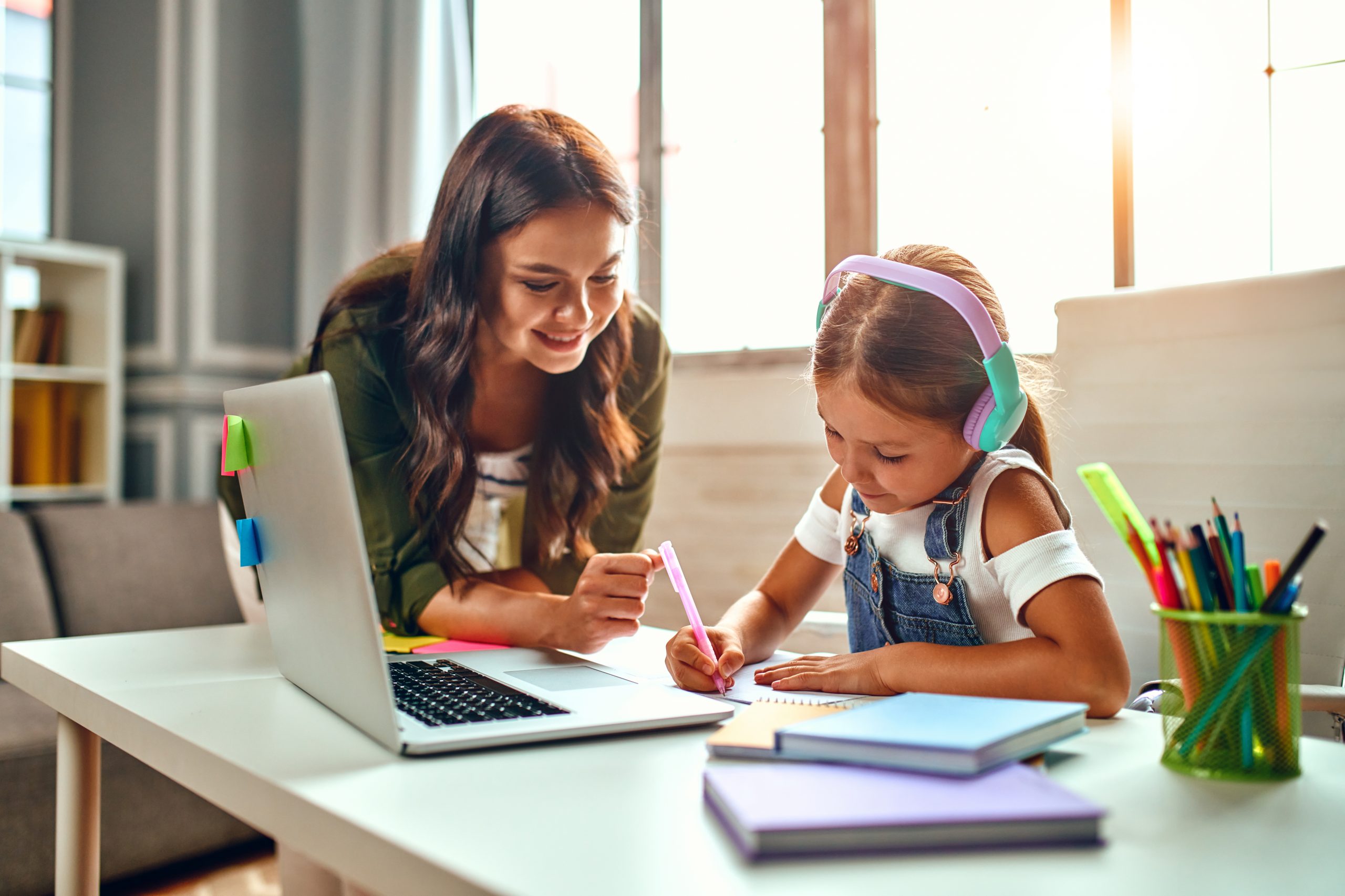 A woman smiles while helping a young girl with headphones as she writes in a notebook, nurturing her ability to cope with change. They sit at a table with a laptop, books, and colorful pencils as sunlight streams through the window, ready to change the world together.