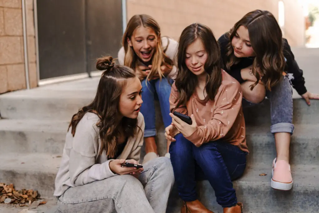 Four girls sitting on outdoor steps are looking at smartphones, engaged and smiling, as they discover how to embrace diversity. Two girls sit directly on the steps while the other two chat just behind them, exploring ways to talk about disabilities with their friends.