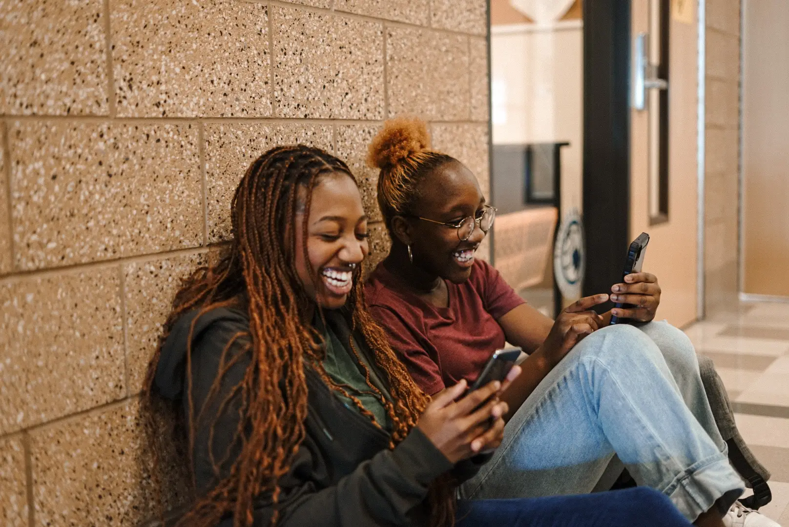 Two friends sit on the floor against a brick wall, smiling and looking at their phones. Engaged in a lighthearted debate about SMS vs MMS, they appear to be enjoying the humorous moment. One wears glasses, and both are casually dressed in comfortable clothes.