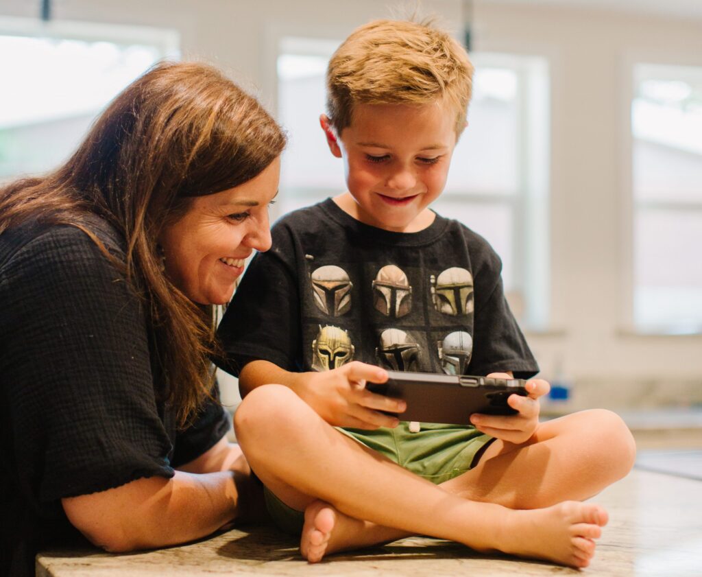 A woman smiles at a young boy immersed in his handheld game console, a perfect alternative to kids wasting the weekend on their phones. The boy sits cross-legged on a table in a bright room with large windows, focused and content.