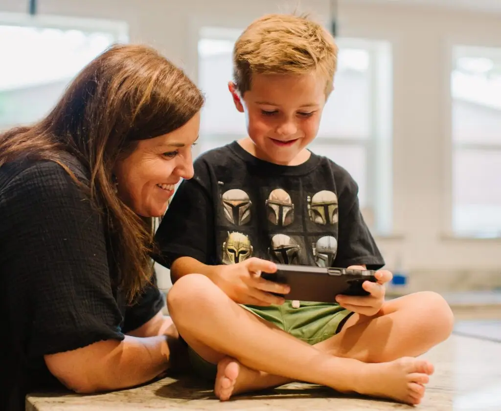 A woman smiles at a young boy immersed in his handheld game console, a perfect alternative to kids wasting the weekend on their phones. The boy sits cross-legged on a table in a bright room with large windows, focused and content.