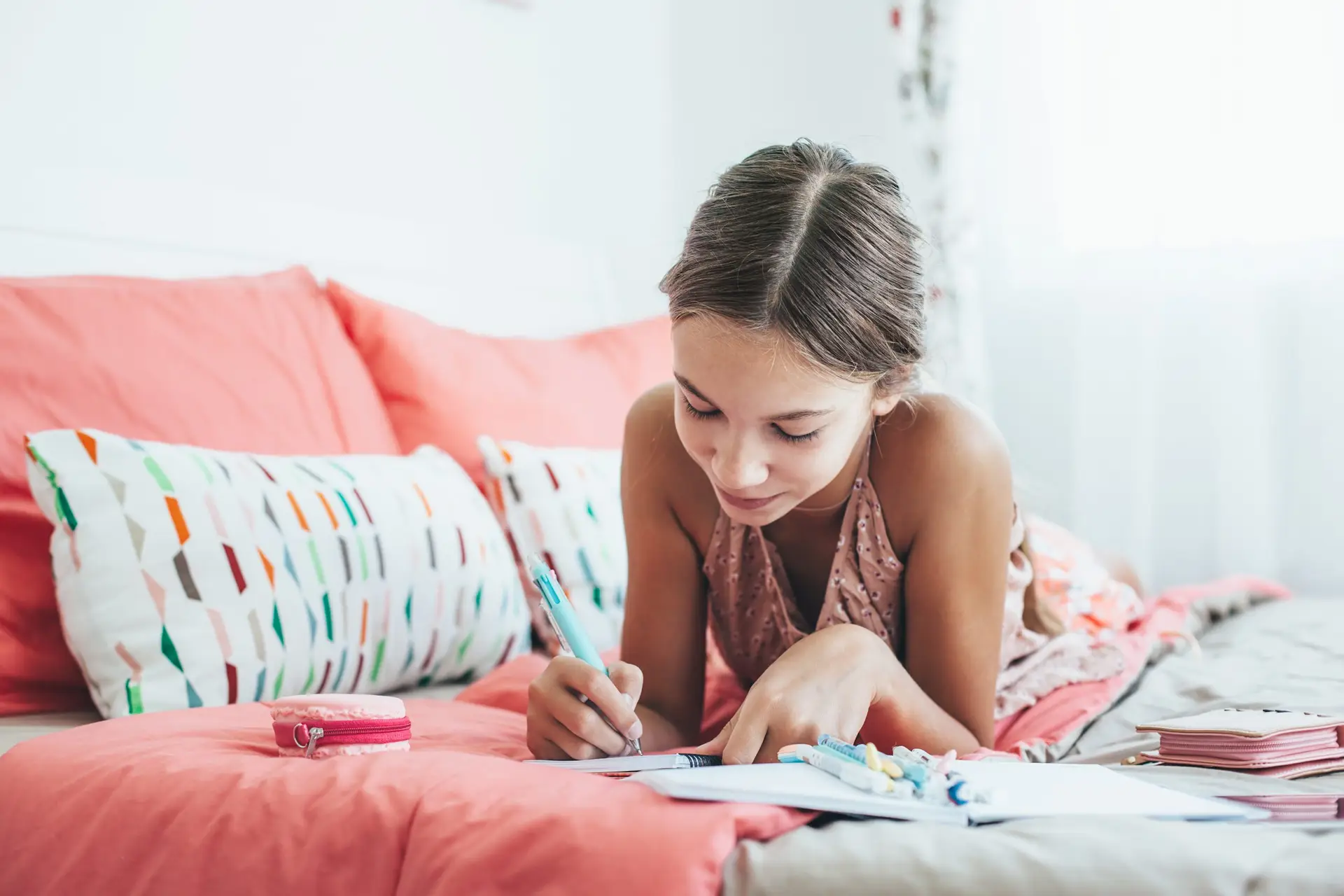 A young girl lies on a bed with coral-colored sheets, diligently writing "5 Tips to Keep Kids Healthy this School Year" in her notebook. Surrounded by patterned pillows and a pink pencil case, she basks in the bright light of her cheerful room.