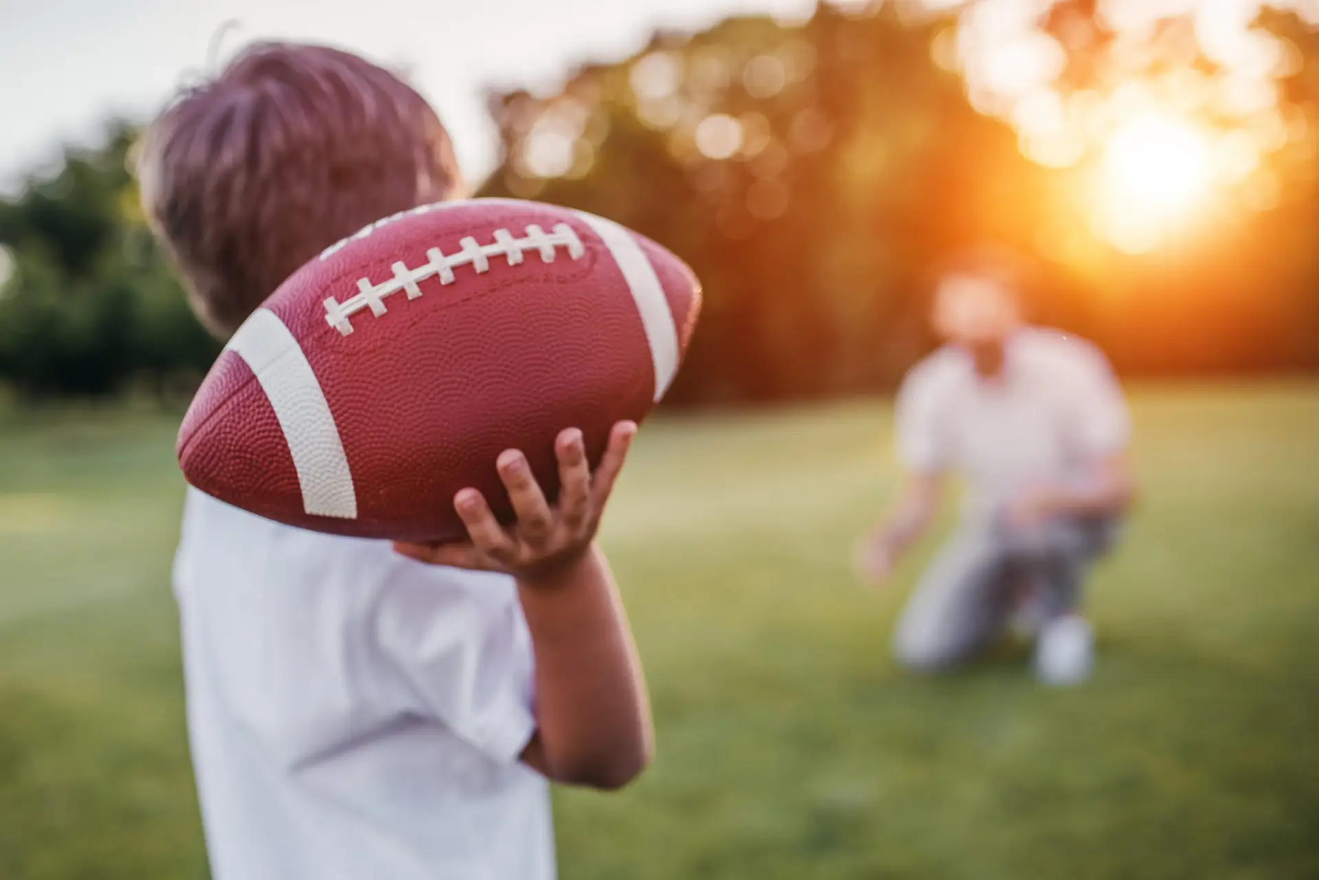 A child holds a football, ready to throw it to an adult in the background. They're playing on a grassy field during sunset, where the benefits of healthy boundaries with your children shine through, as sunlight creates a warm glow in the scene.