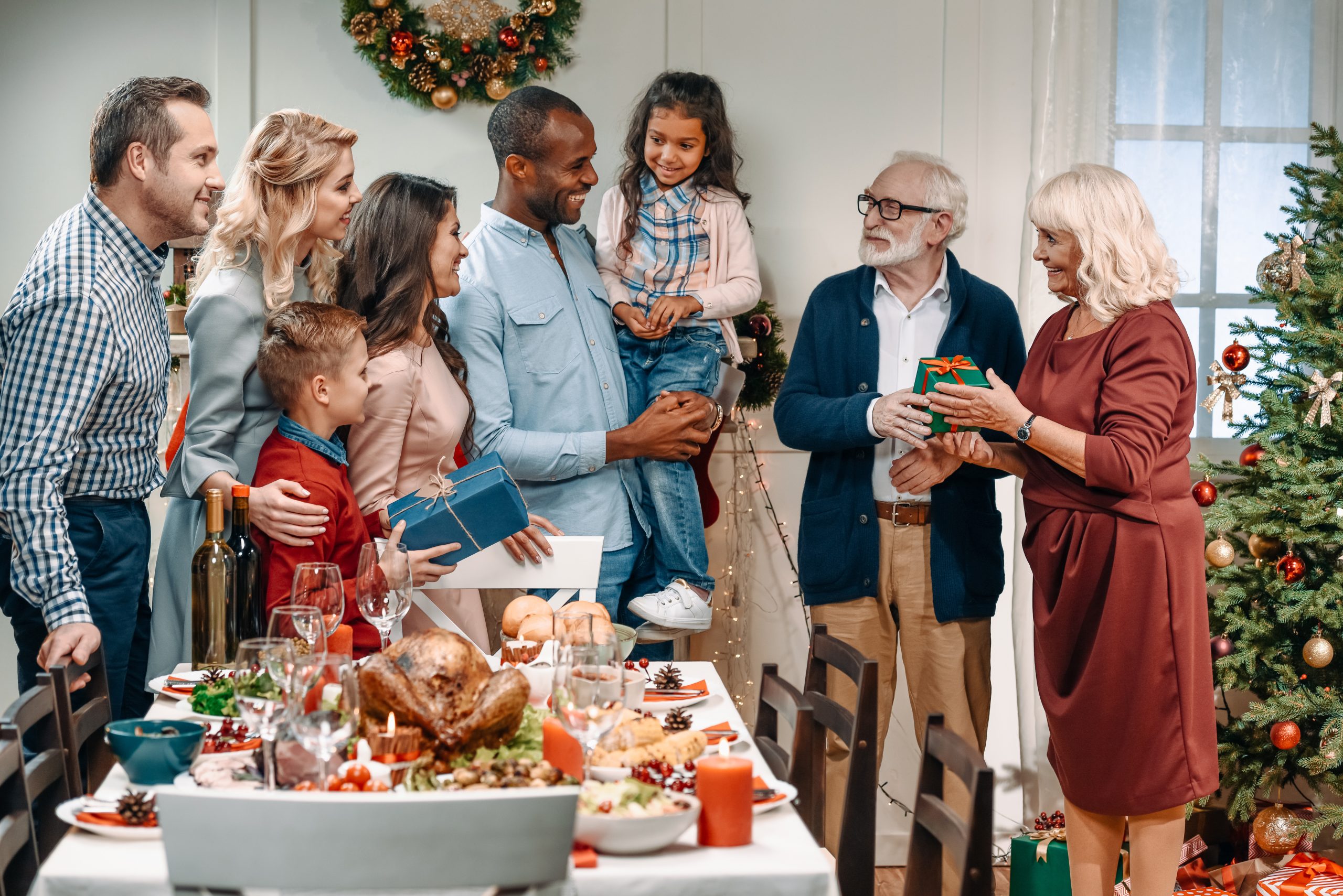 A group of people gather around a festive table set with food and drinks. An elderly woman hands a gift to an elderly man, embodying the importance of Christmas traditions for kids. A young child smiles while being held by an adult, as a Christmas tree and wreath decorate the room.