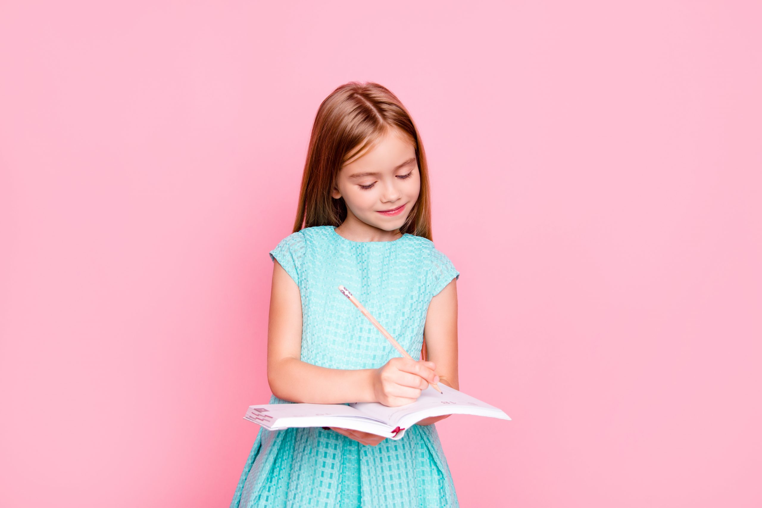 A young girl with long brown hair in a blue dress is planning out her day, writing thoughtfully in a notebook with a pencil. She stands against a solid pink background, smiling and focused on whether paper or digital choices suit her best.