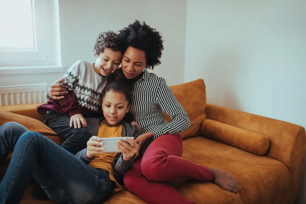 A mother and her two children are sitting on a mustard-colored couch. The older child is exploring a smartphone glossary, while the younger child leans affectionately against the mother's shoulder. They are all smiling and appear to be enjoying a moment together.