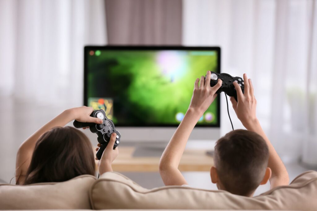 Two children are sitting on a couch, playing the latest video game on a console. They each hold a controller, and the TV screen in front of them shows a brightly colored game scene. The softly lit room is filled with natural light, ensuring they enjoy new games while keeping kids safe indoors.