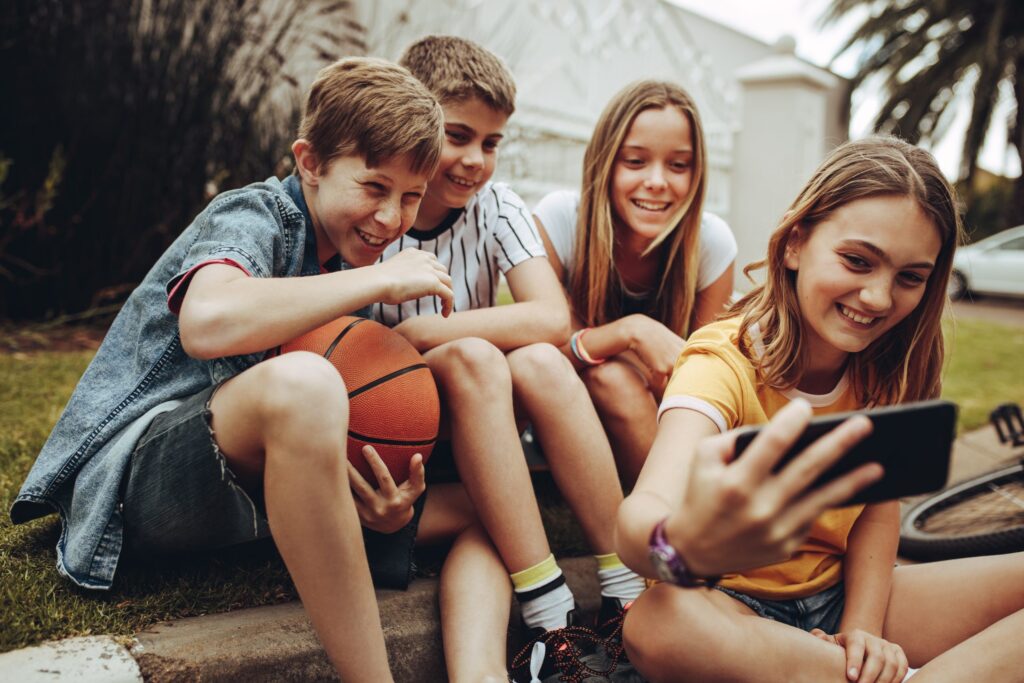 Four children sit on the grass, taking a selfie with their Troomi phone. One holds a basketball, and they all appear happy, smiling at the device. It's a sunny day, with a house and palm trees in the background—proof that fun doesn't need social media.