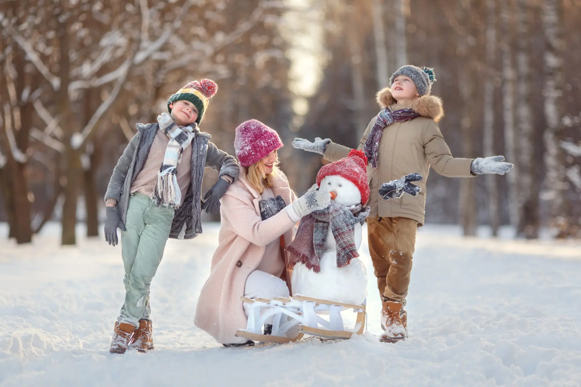 A woman kneels in the snow with two children, all smiling and savoring fun, stress-free snow day activities for the whole family. Dressed in winter clothes, they stand beside a cheerful snowman with a red hat and scarf against a backdrop of snow-covered trees and a sled.