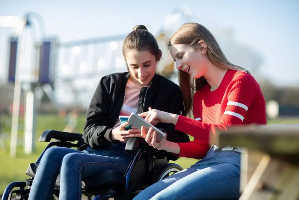 Two young women are outdoors at a park, delighting in the wonders of assistive technology tools. One sits in a wheelchair as they both smile and engage with a smartphone. In the blurred background, a playground invites possibilities with tech for kids with disabilities.