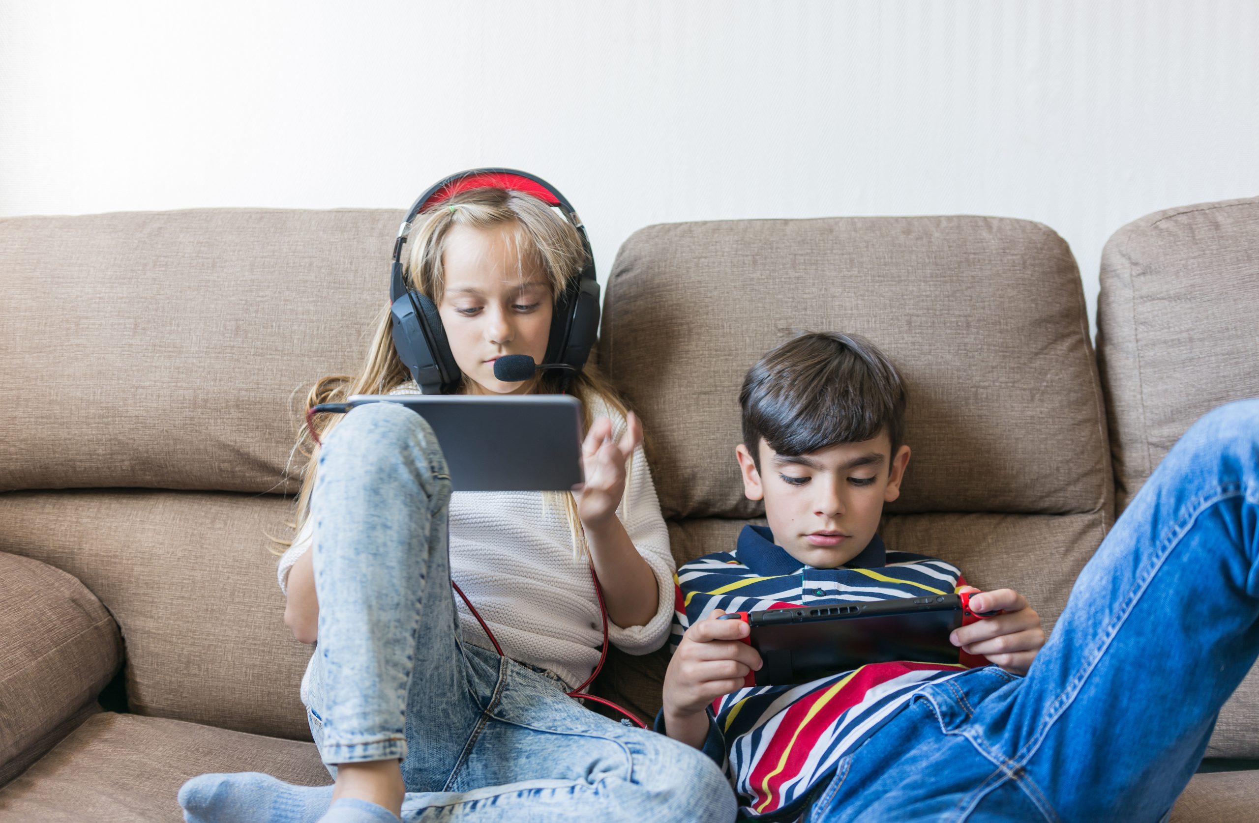 Two children sit on a couch, showcasing some of the latest inventions to make kids' lives easier. The girl on the left wears headphones and uses a tablet, while the boy on the right is engrossed in his handheld gaming device. Both are casually dressed in jeans and T-shirts.