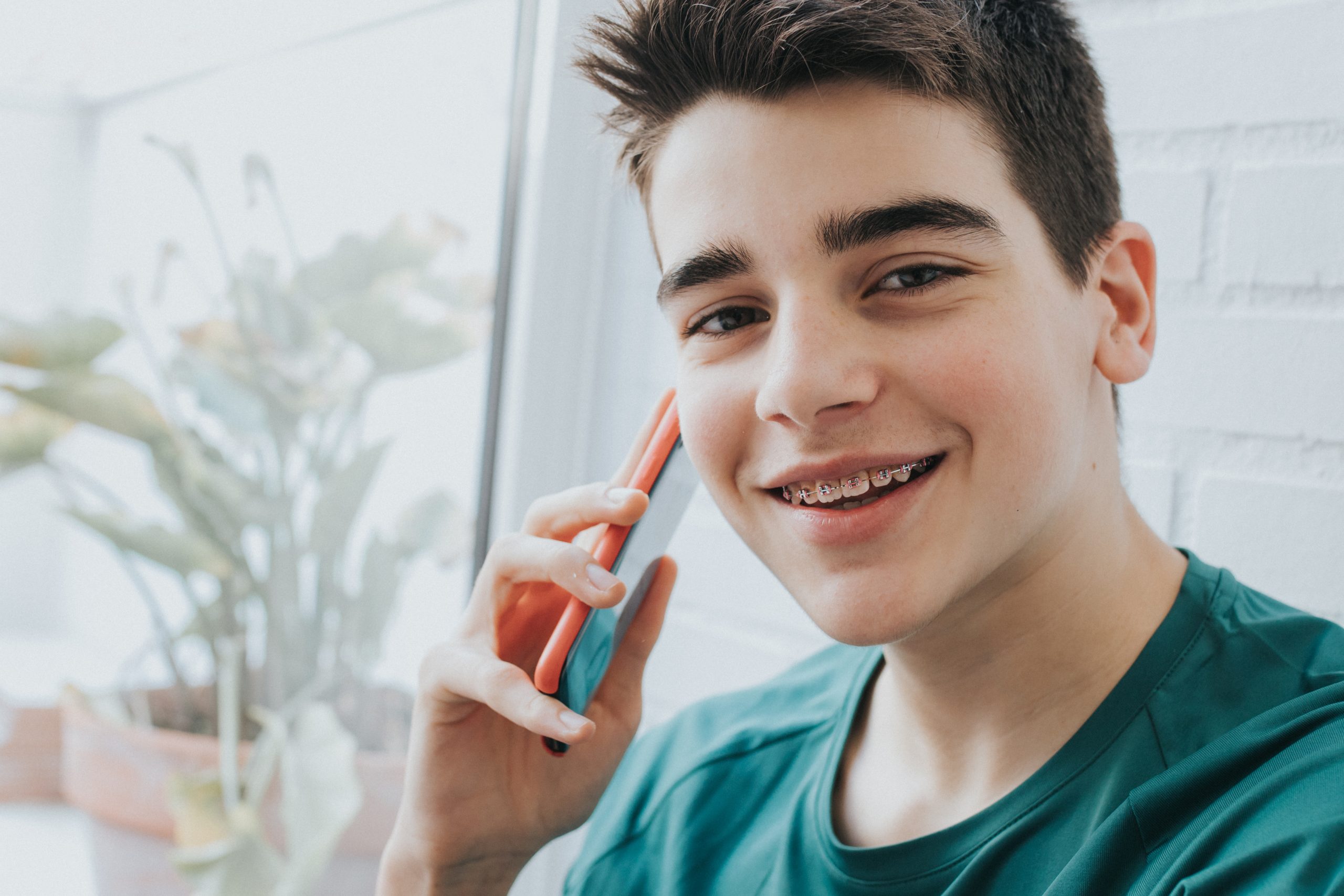 A smiling teenager with braces holds a colorful Troomi phone to his ear. He is sitting near a window, where the only spam he's worried about is on his sandwich, set against a blurred background of potted plants.